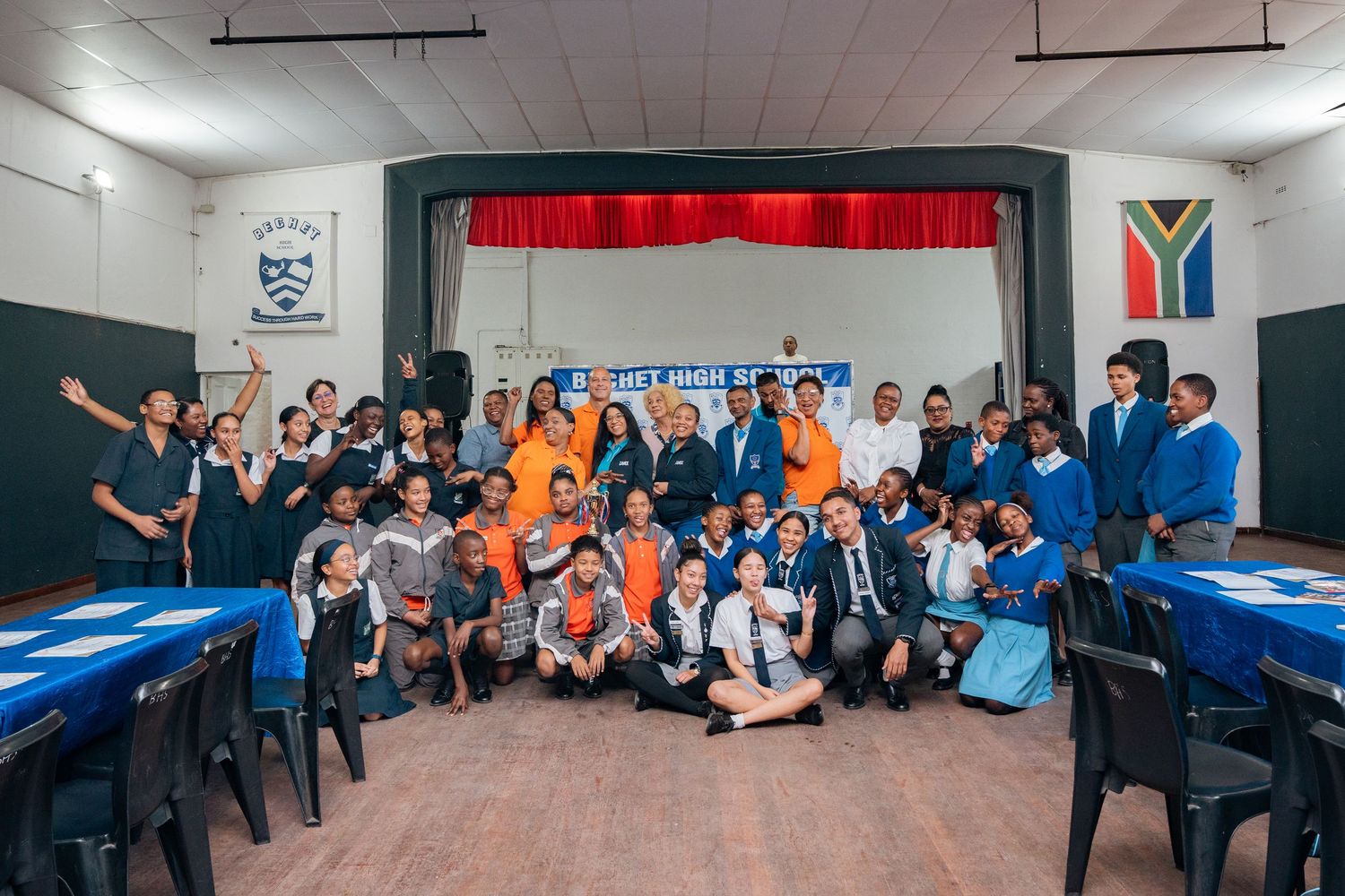 Large group of students and teachers posing together in a school hall with South African flag displayed.