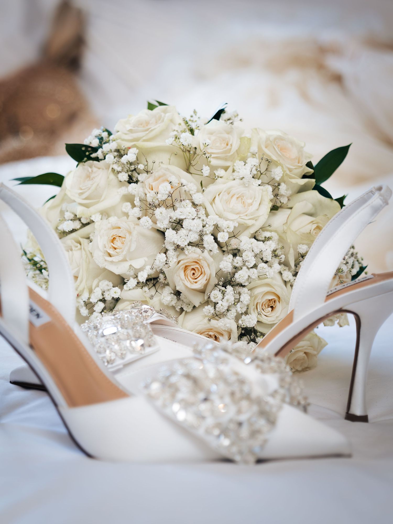 Elegant bridal bouquet of white roses and baby's breath beside crystal-embellished white satin wedding heels.