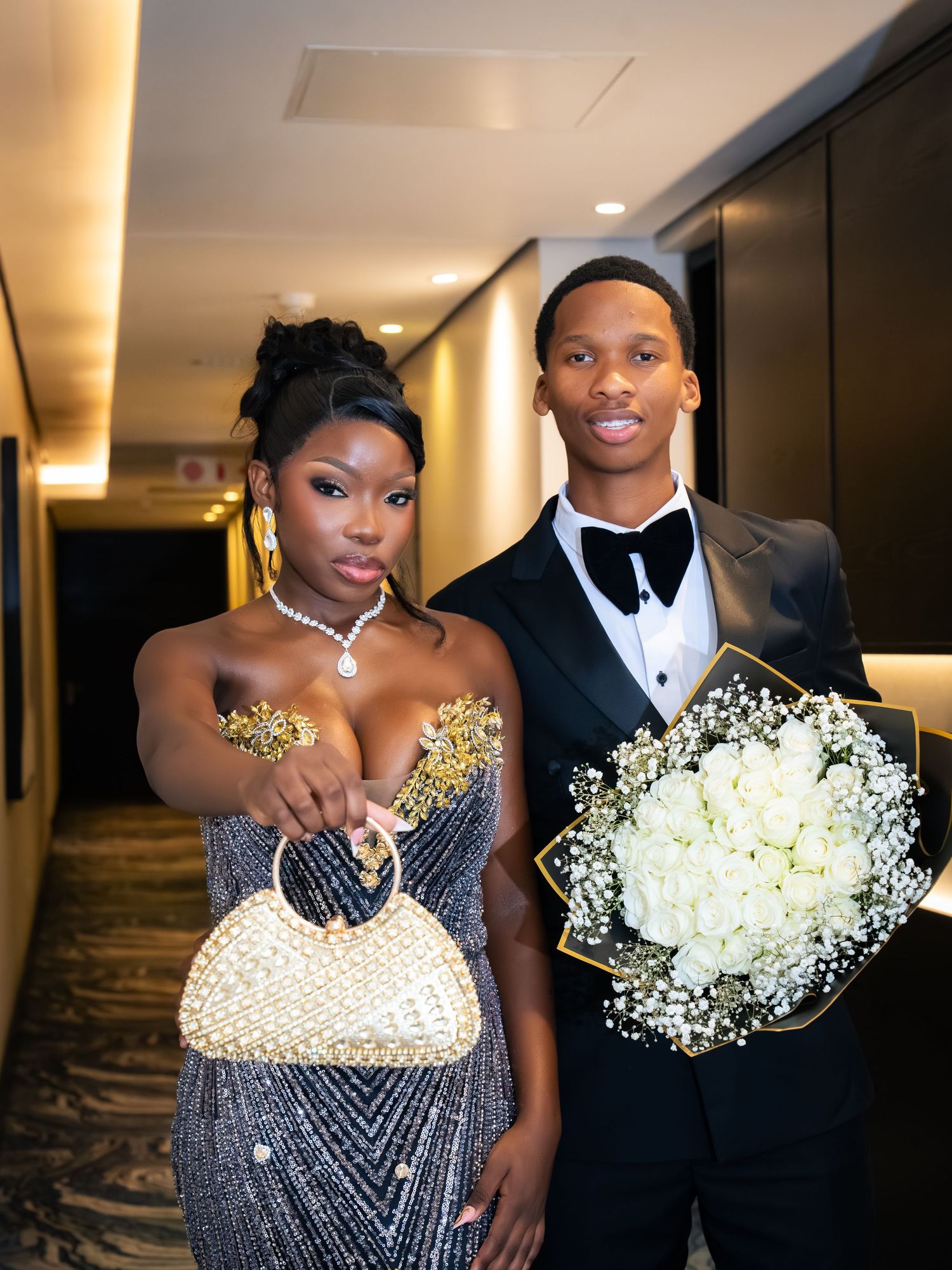 Elegant couple in formal attire, woman in beaded gown with gold accents, man in tuxedo holding white rose bouquet.
