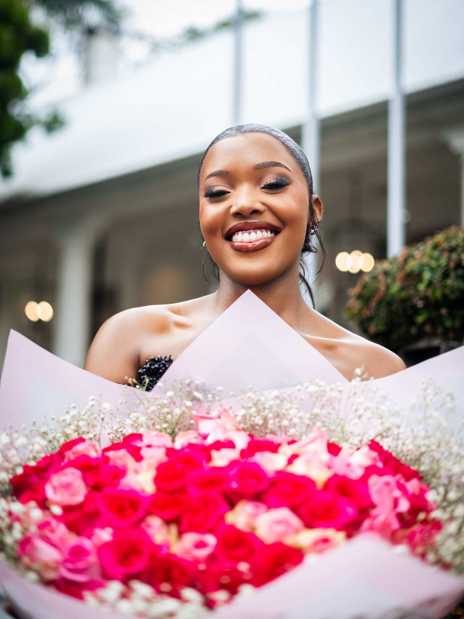 Smiling woman holding a large bouquet of pink and red roses wrapped in white paper outdoors.