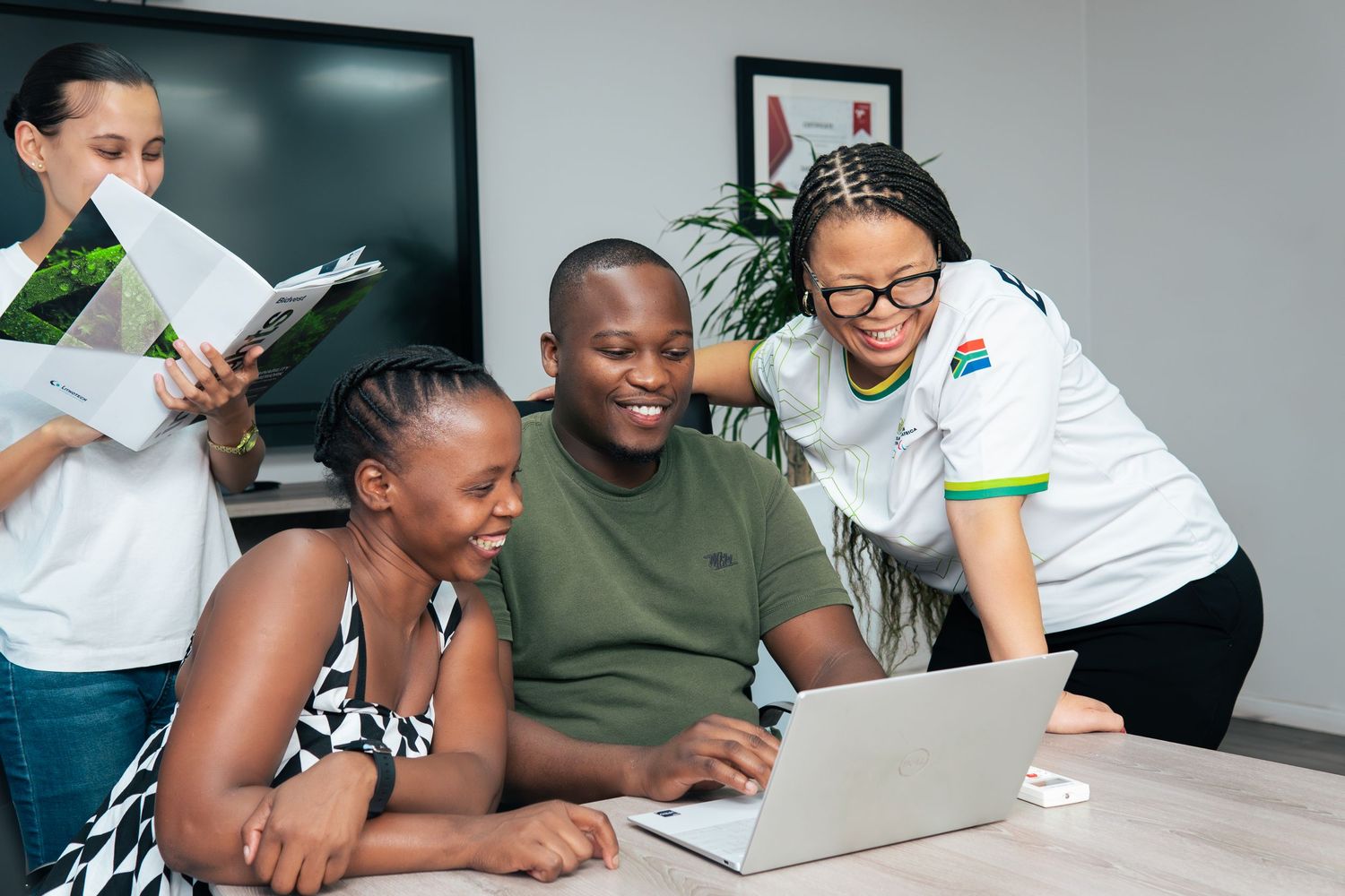 Three colleagues smile while reviewing work on a laptop together in a modern office meeting room.