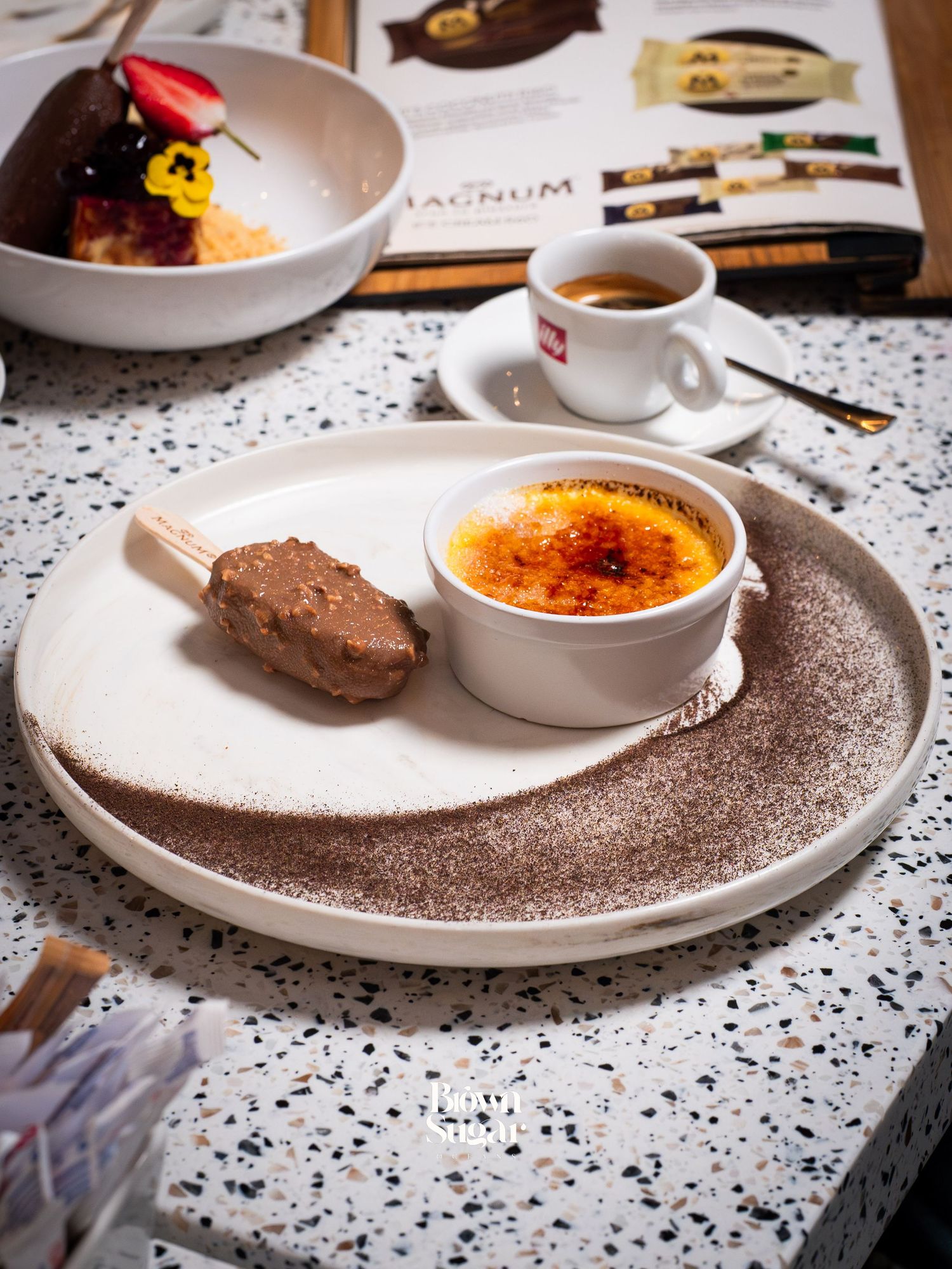 Elegant dessert plate with crème brûlée, chocolate ice cream bar, and espresso coffee cup on terrazzo surface.