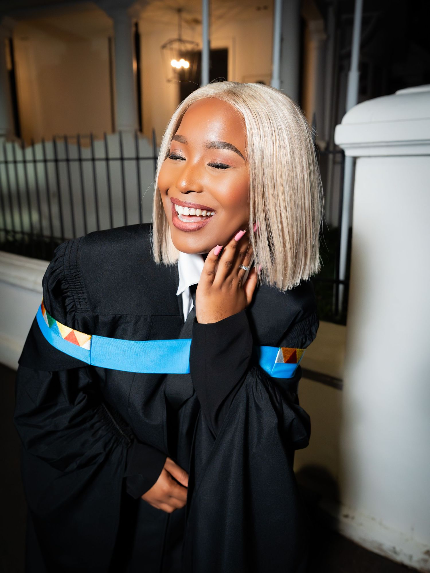 Smiling graduate in black gown with blue sash, blonde bob hairstyle, celebrating outdoors.