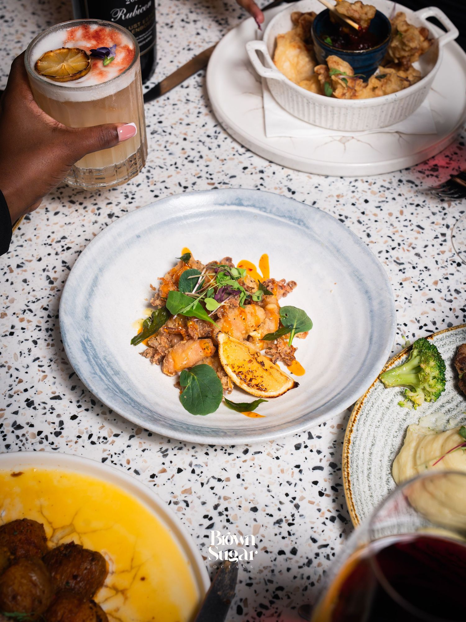 Elegant restaurant table with shrimp dish garnished with greens, seafood casserole, and cocktails on terrazzo surface.