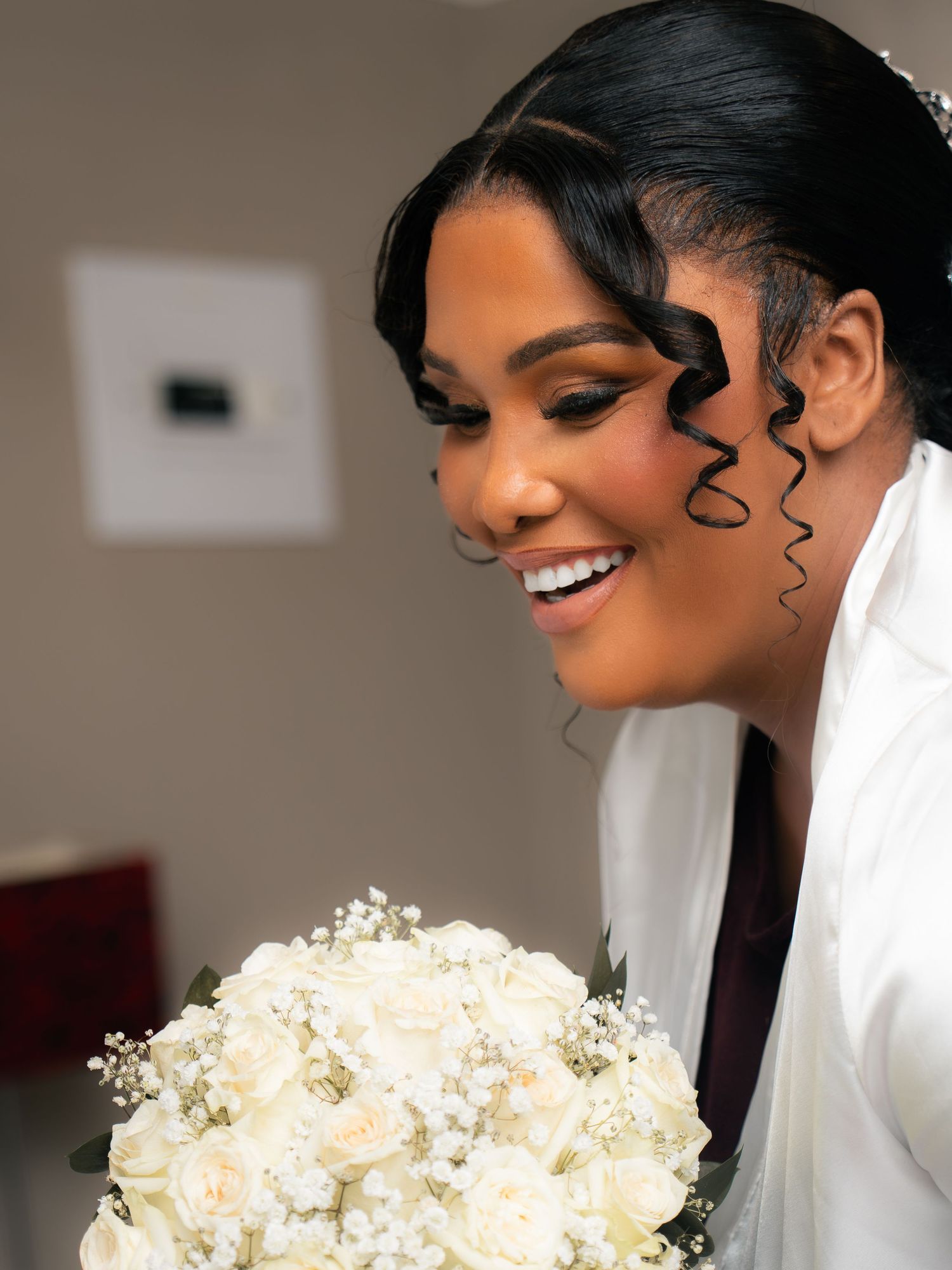 Smiling bride holding white rose bouquet with elegant curled hair tendrils and glam makeup on her wedding day.