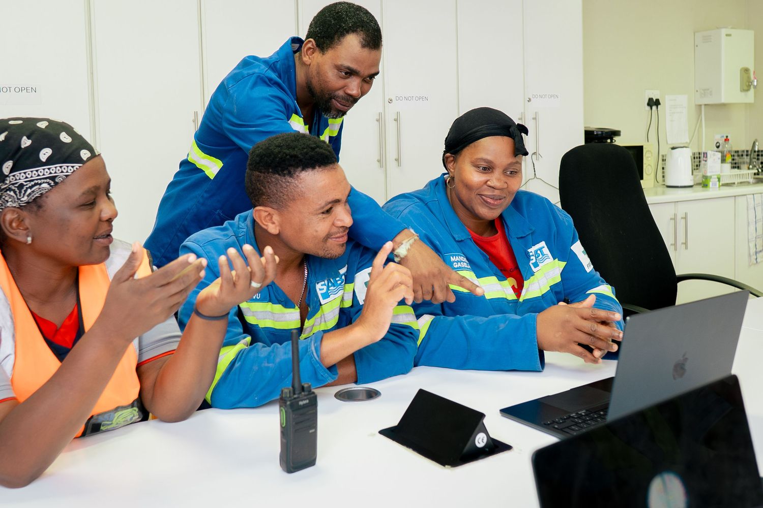 Four workers in blue uniforms collaborate enthusiastically around a laptop in a bright office setting.