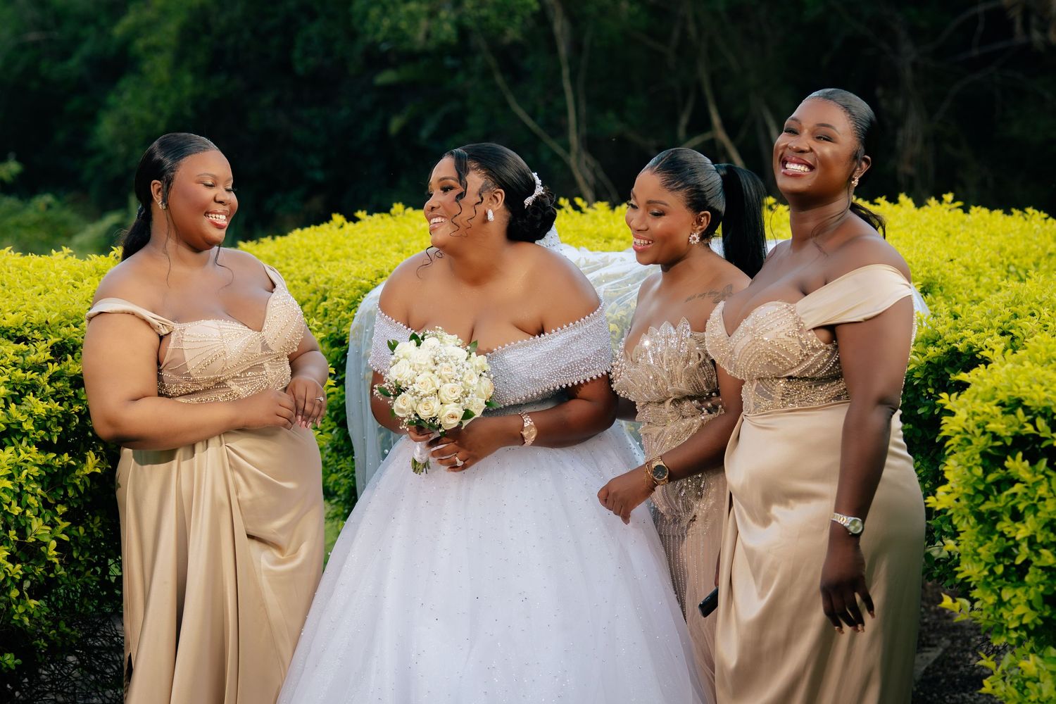 Bride in white gown with bridesmaids in gold dresses holding bouquet outdoors at wedding celebration.