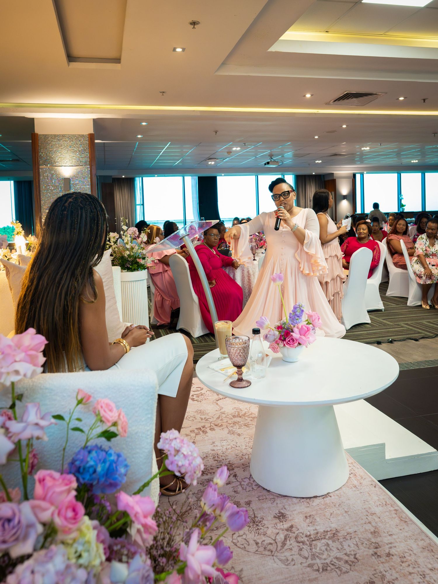 Elegant event venue with guests in pink attire, floral arrangements, and a speaker presenting at a styled white table.