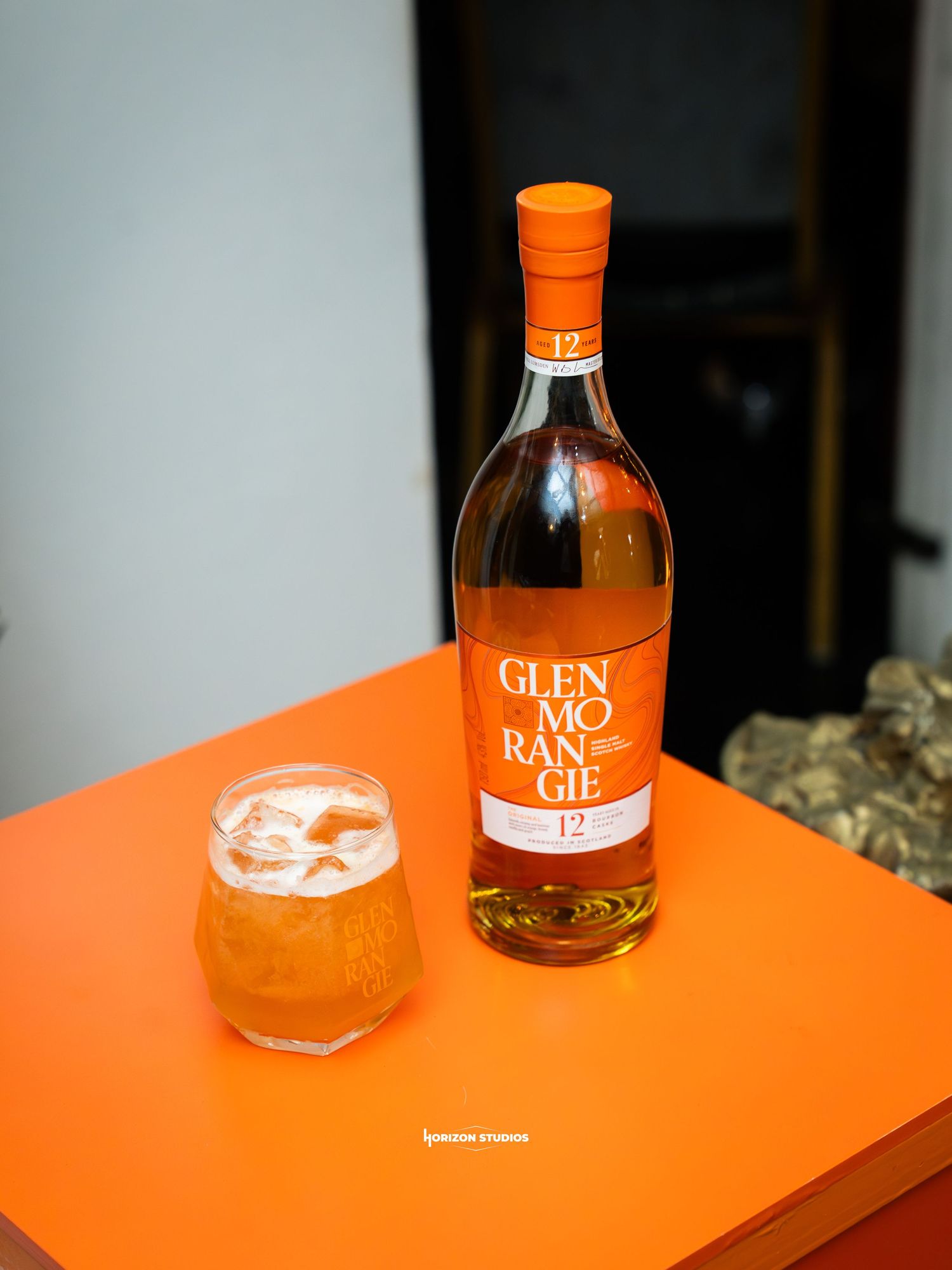 Glenmorangie 12 year old whisky bottle beside a cocktail glass on an orange table.