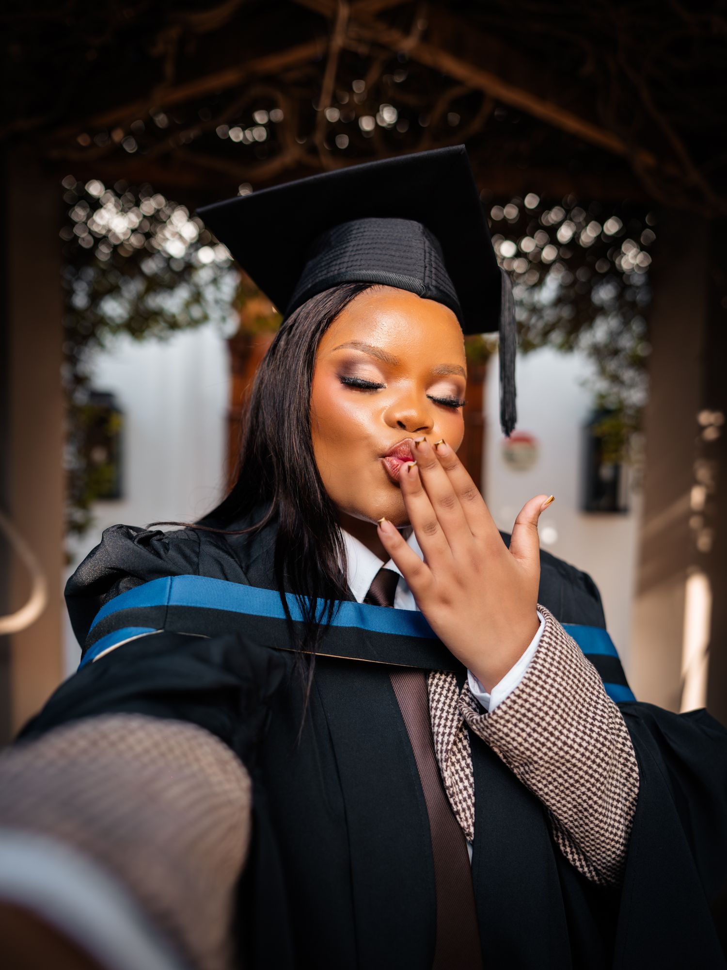A young woman in graduation cap and gown blows a kiss during her graduation photo session outdoors.