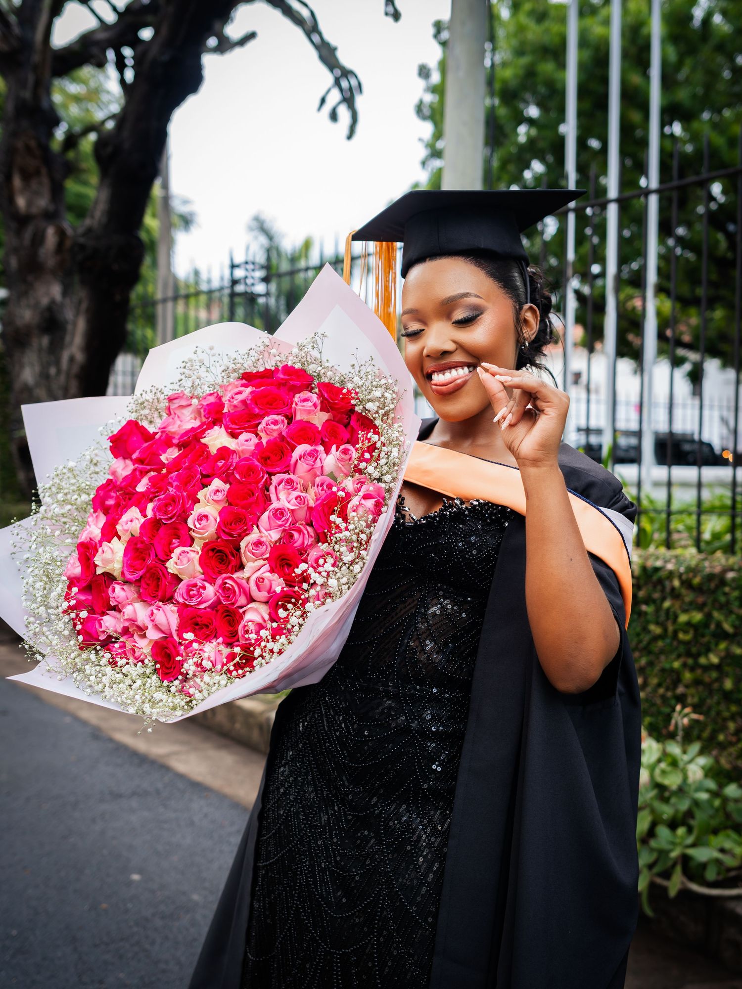 Smiling graduate in cap and gown holding large pink rose bouquet with baby's breath outdoors.
