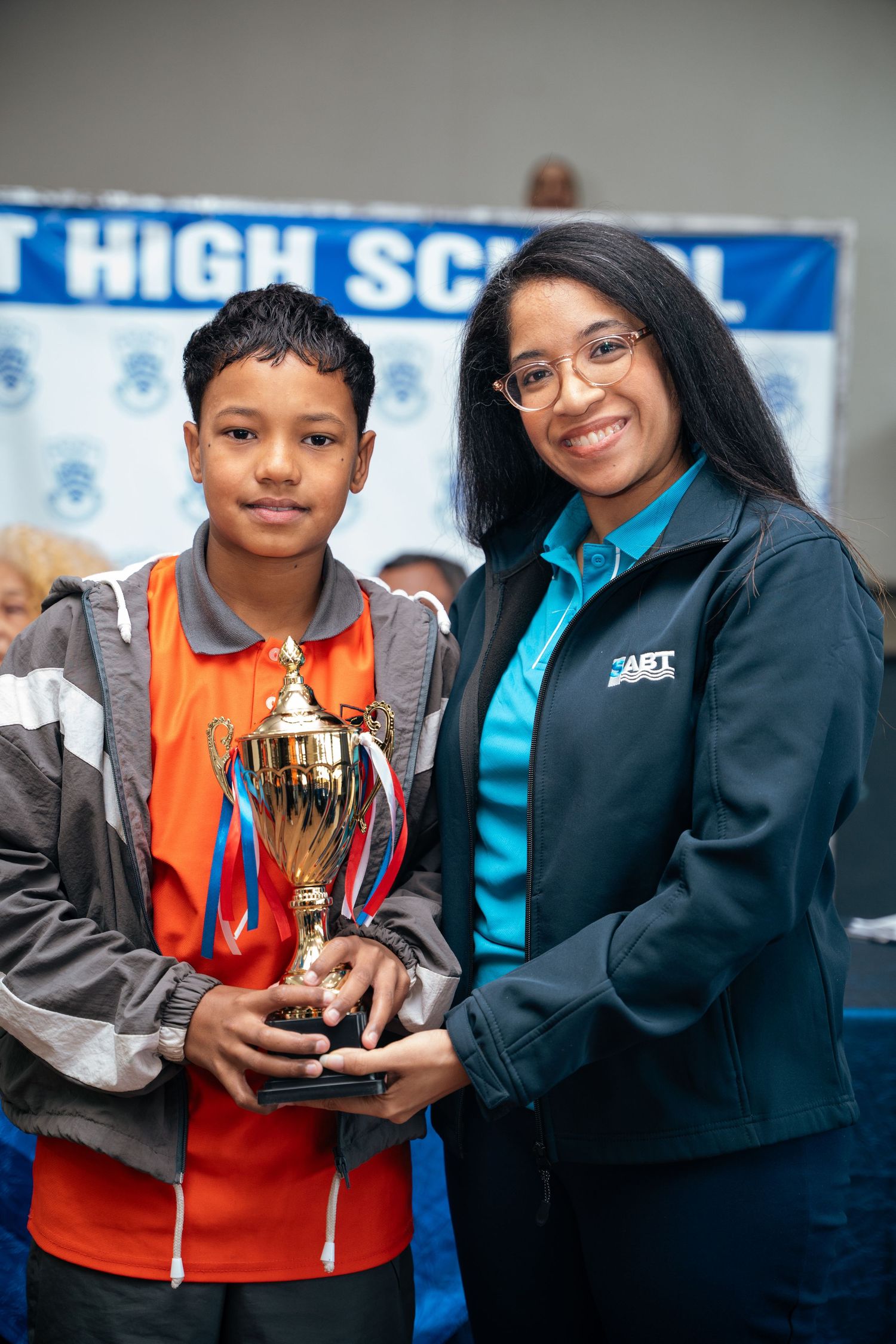 A young student and a teacher proudly hold a gold trophy together in front of a high school banner.