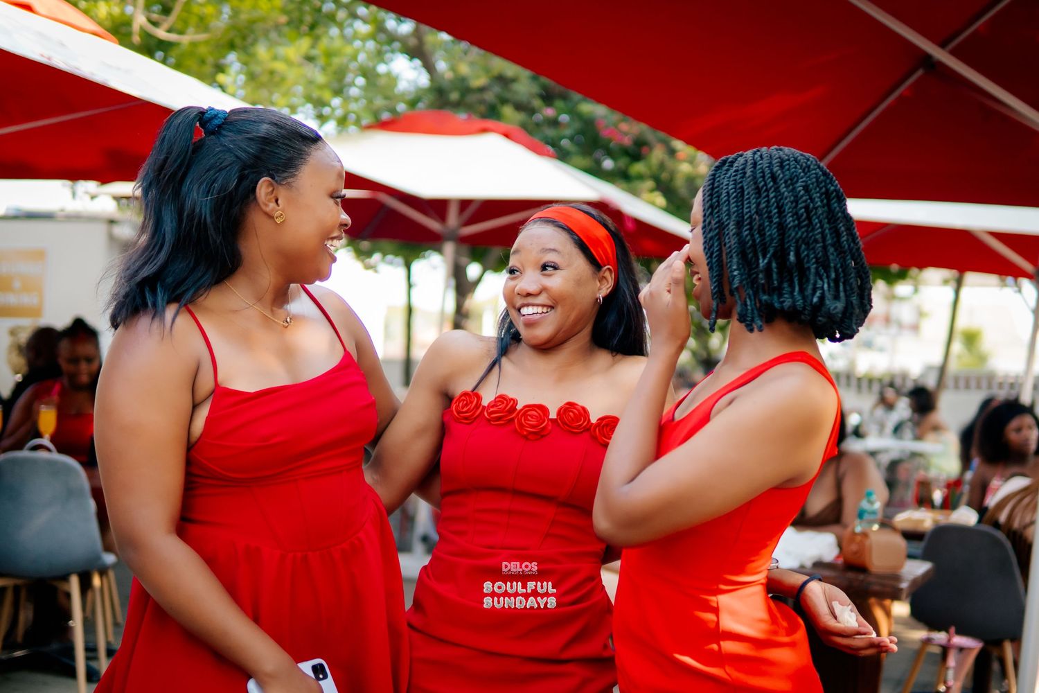 Three women in red dresses laugh together at an outdoor social event.