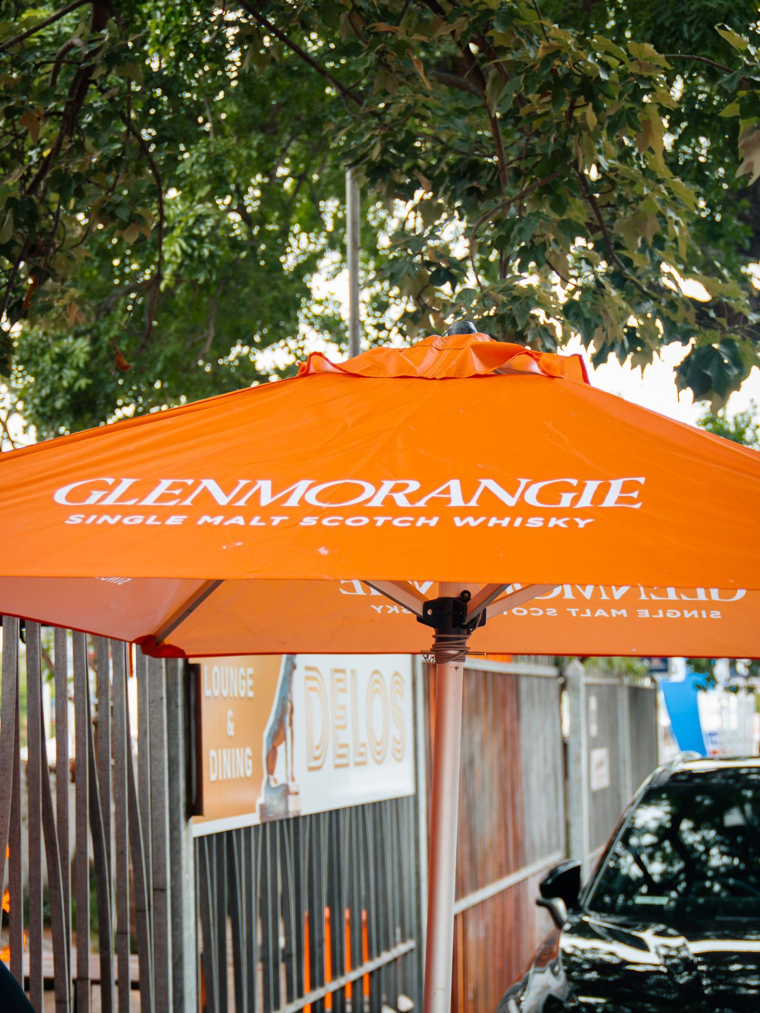 Orange Glenmorangie Single Malt Scotch Whisky branded patio umbrella outdoors under green trees.
