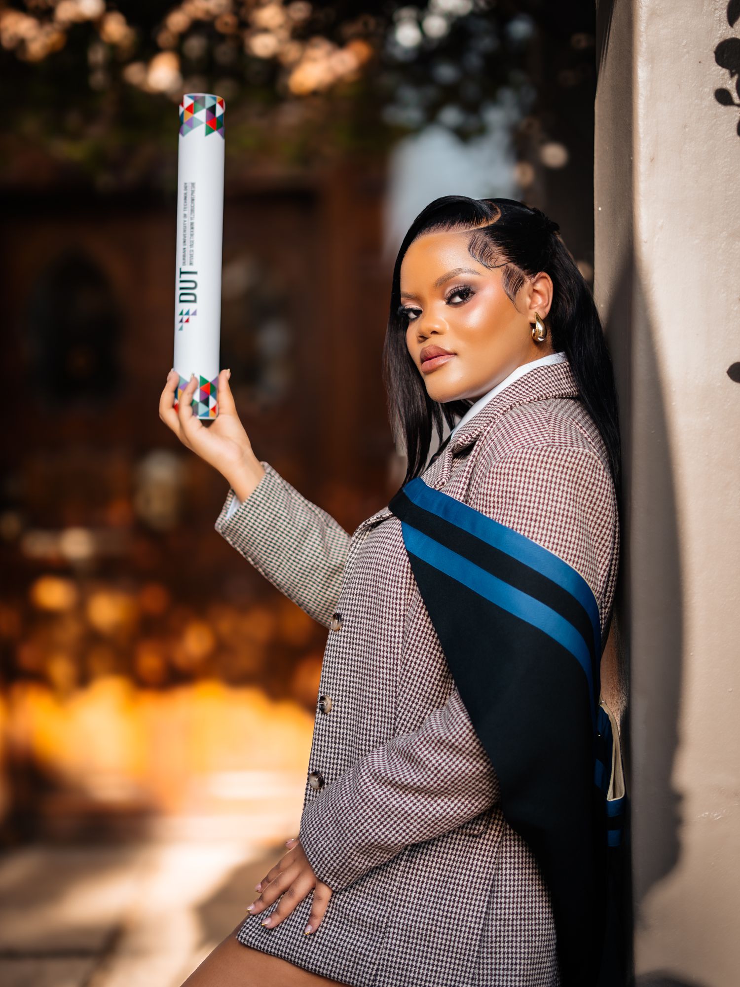 Young woman in plaid blazer poses outdoors holding a diploma tube with a blue sash, celebrating graduation.