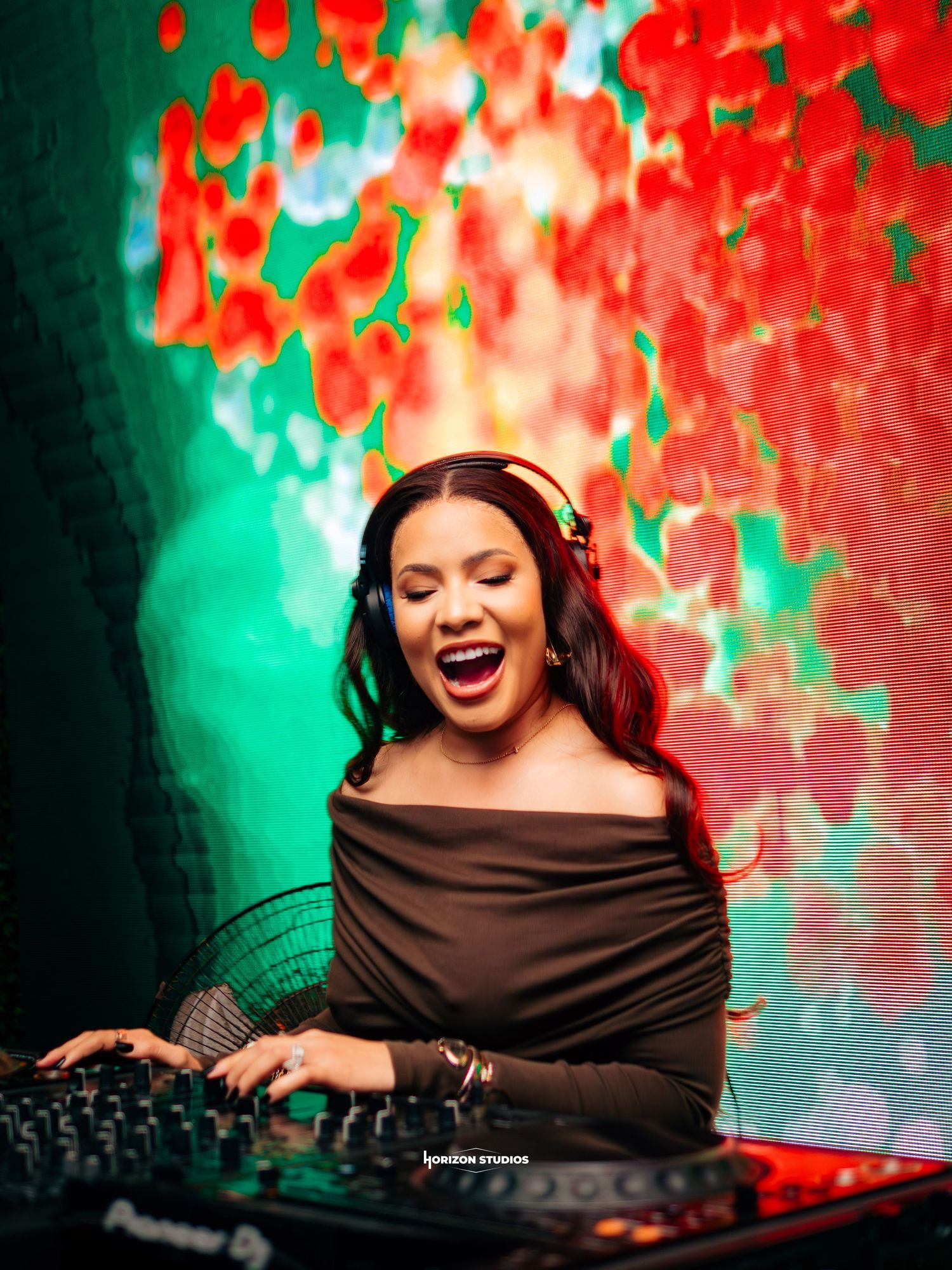 A female DJ laughs joyfully while mixing at Pioneer decks, wearing headphones and an off-shoulder top against a vibrant floral backdrop.