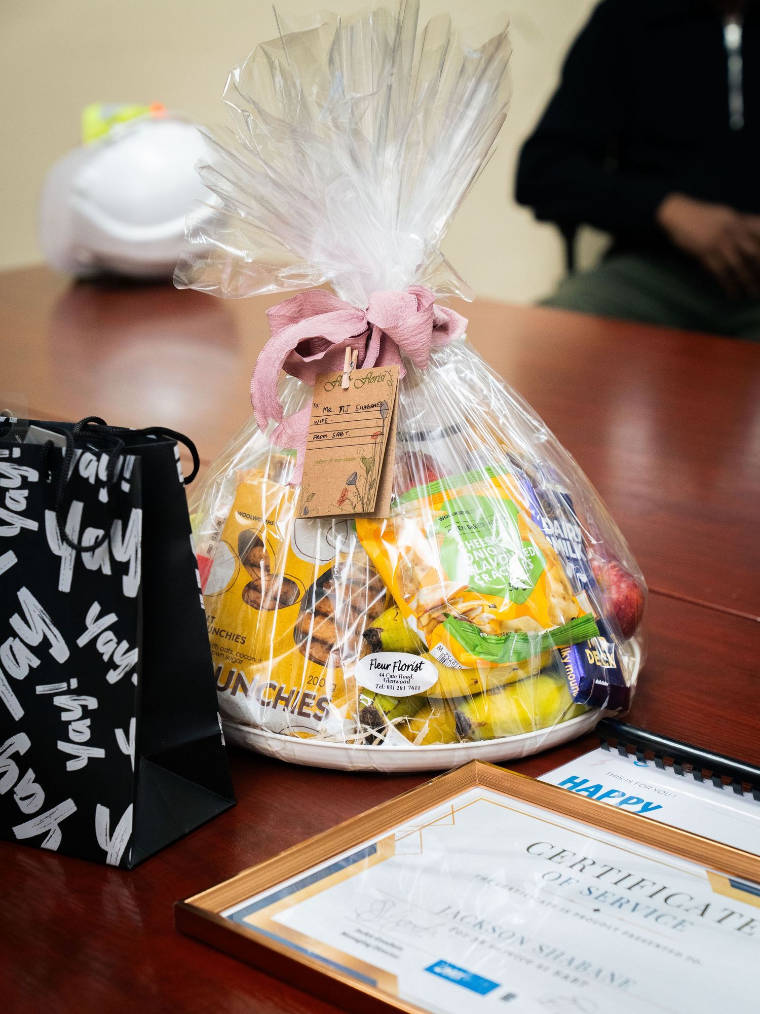 A gift basket filled with snacks and candy, tied with a pink bow, displayed on a table with a certificate and gift bag.