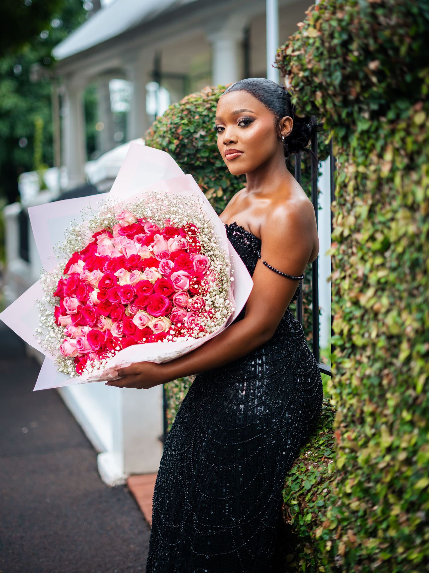 Elegant young Black woman in black sequin gown holding large pink rose bouquet outdoors.