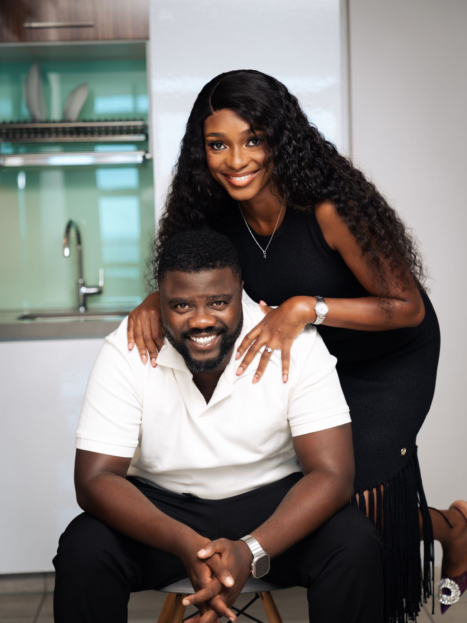 A smiling couple poses together in a modern kitchen, woman in black dress stands behind seated man in white shirt.