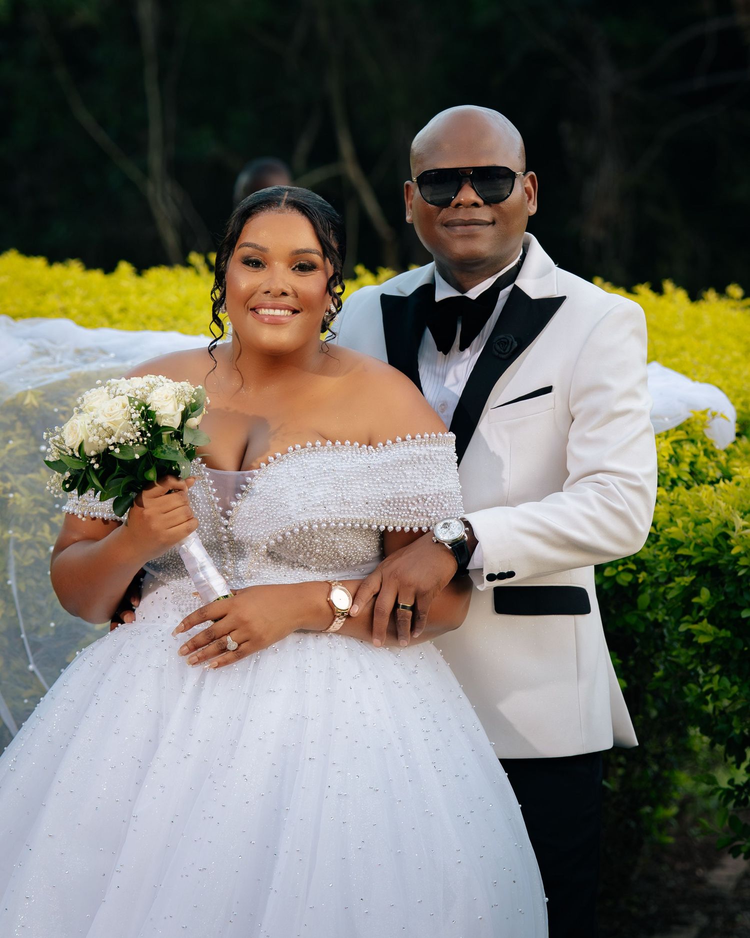 Elegant couple posing at wedding, bride in white ballgown with bouquet, groom in white tuxedo with sunglasses.