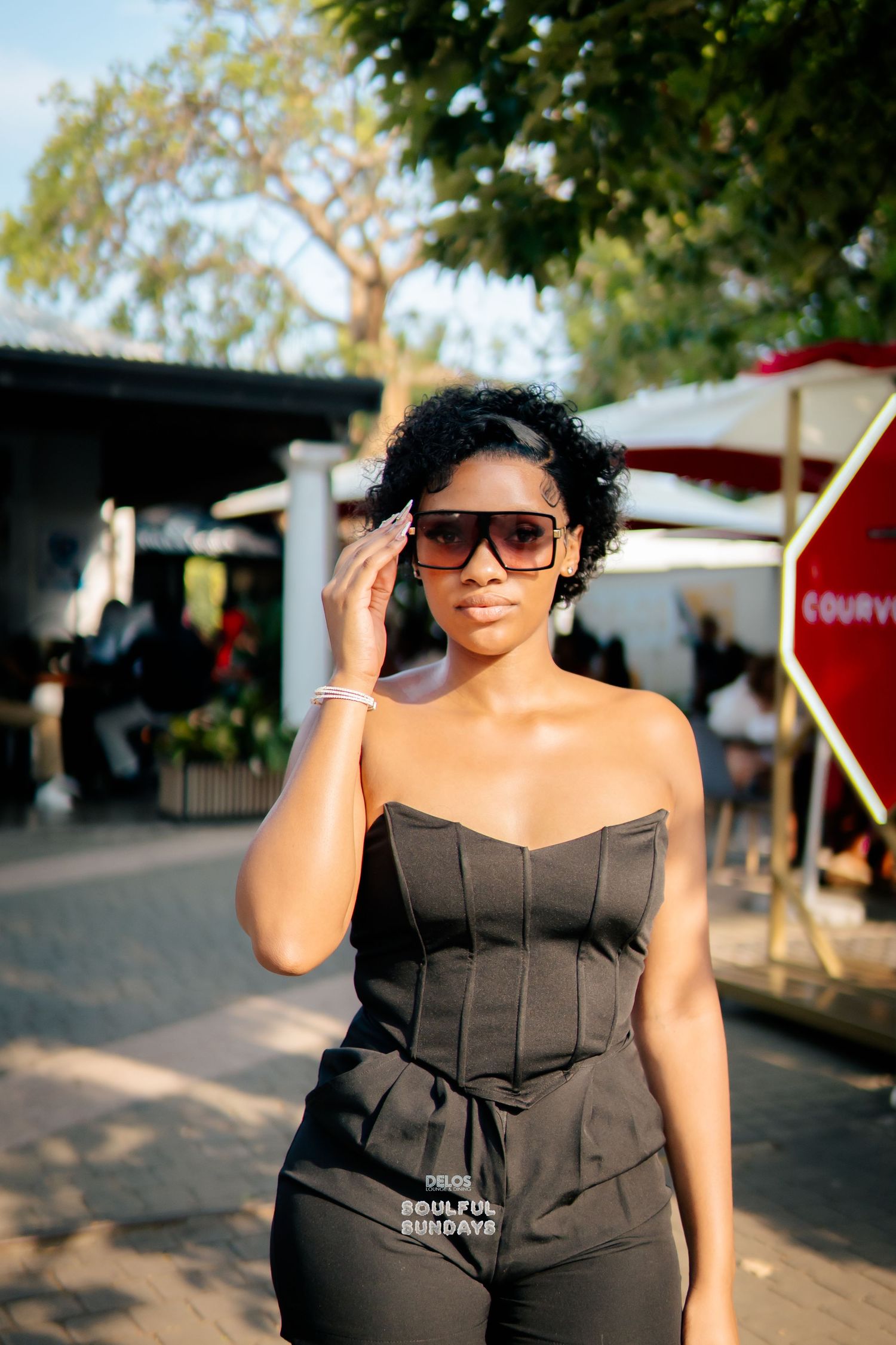 A stylish woman in a black strapless corset outfit wearing oversized sunglasses poses outdoors at a sunny event.