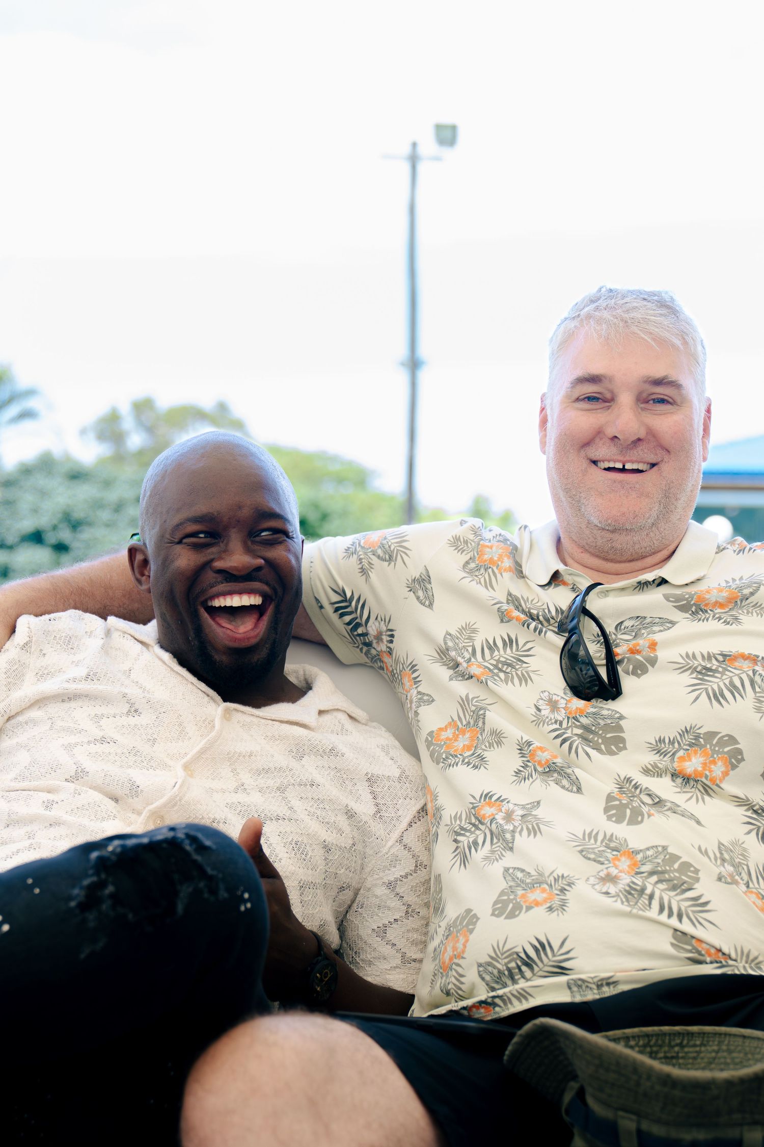 Two smiling men sitting together outdoors, one in a floral shirt with sunglasses, enjoying a cheerful moment.