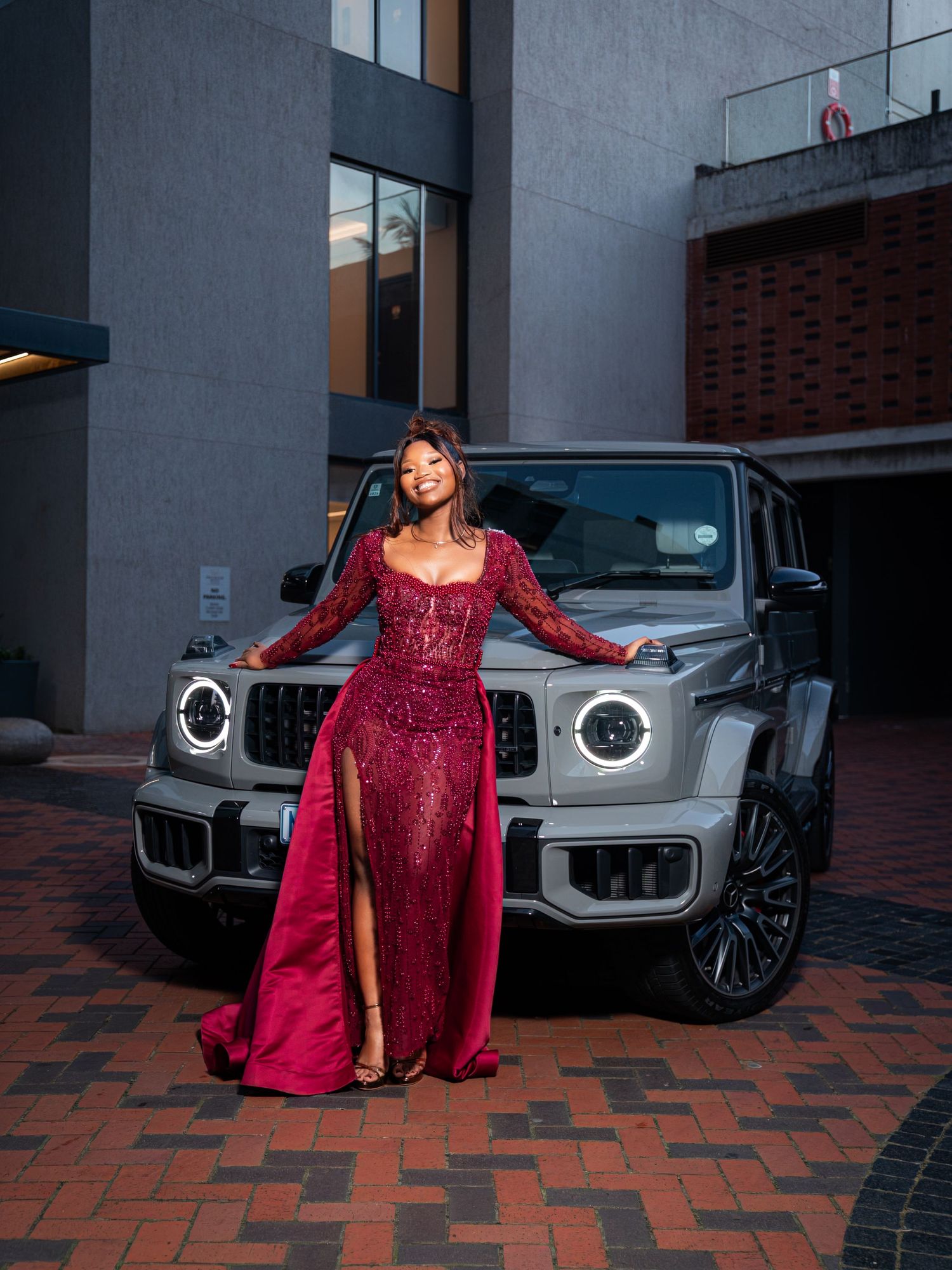 Woman smiling in red sequin gown with high slit standing in front of Mercedes G-Class SUV.