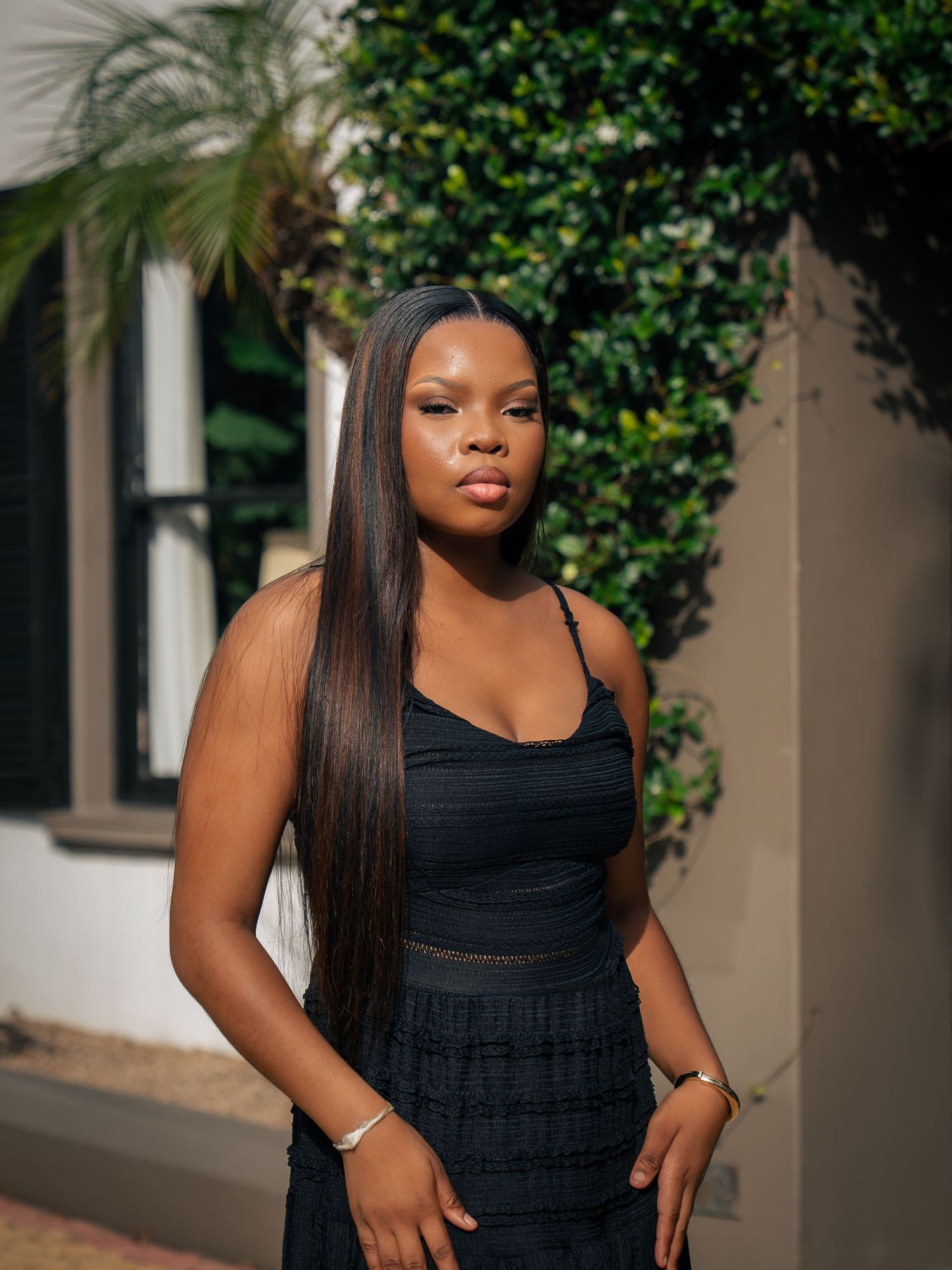A young Black woman in a black outfit with long straight hair poses outdoors near green hedges and a white wall.