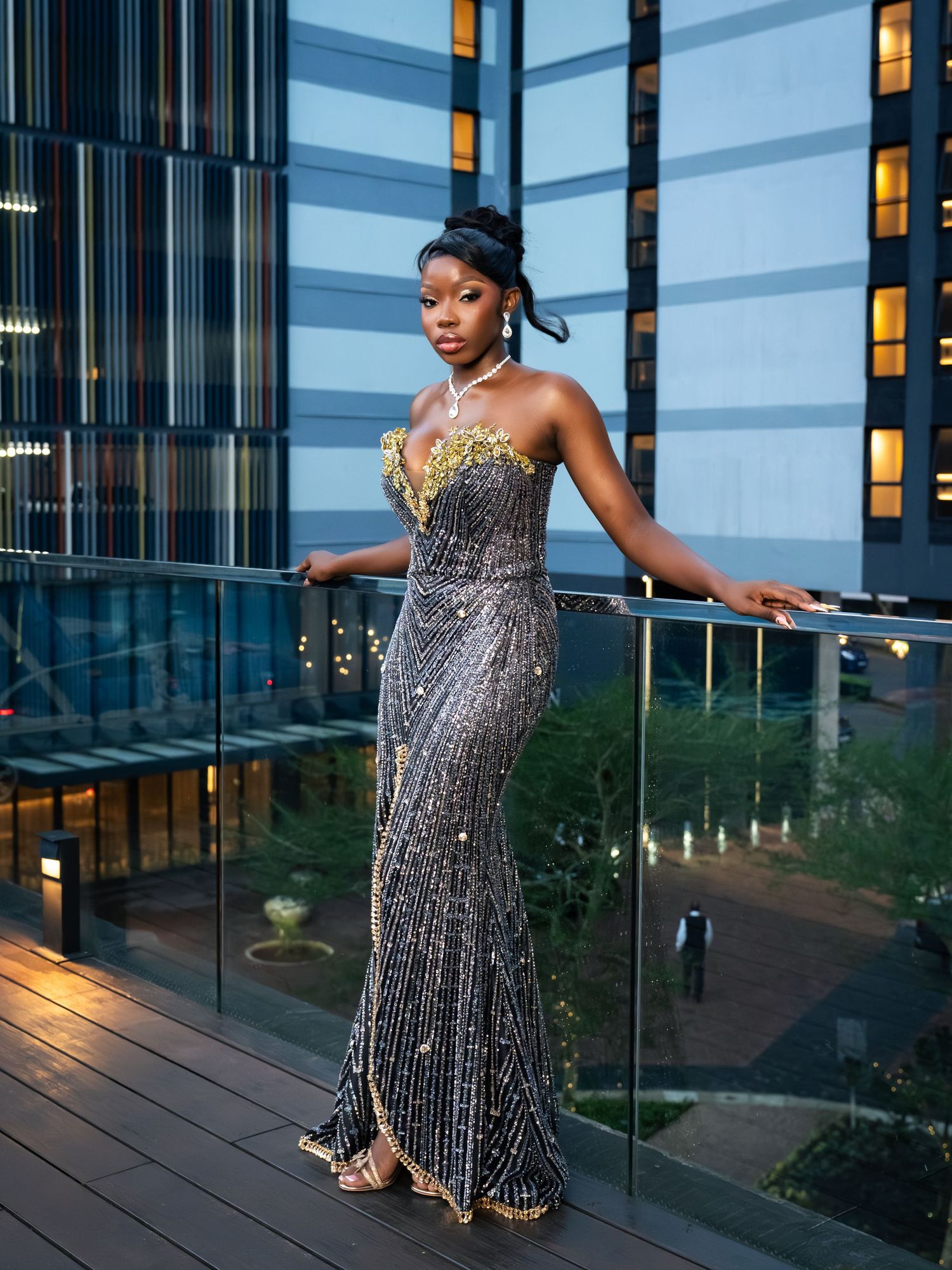 A woman in a stunning beaded gray and gold strapless gown poses elegantly on a modern building balcony at dusk.