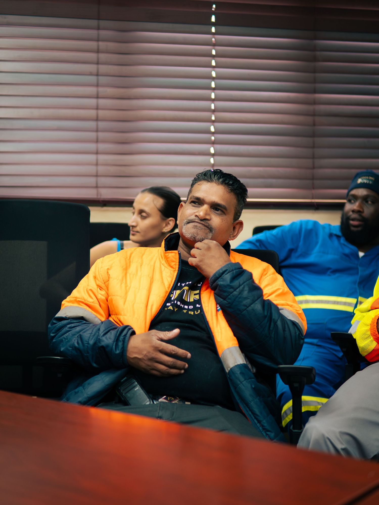 Three workers in high-visibility jackets sit in a meeting room, with one man thoughtfully resting his hand on his chin.