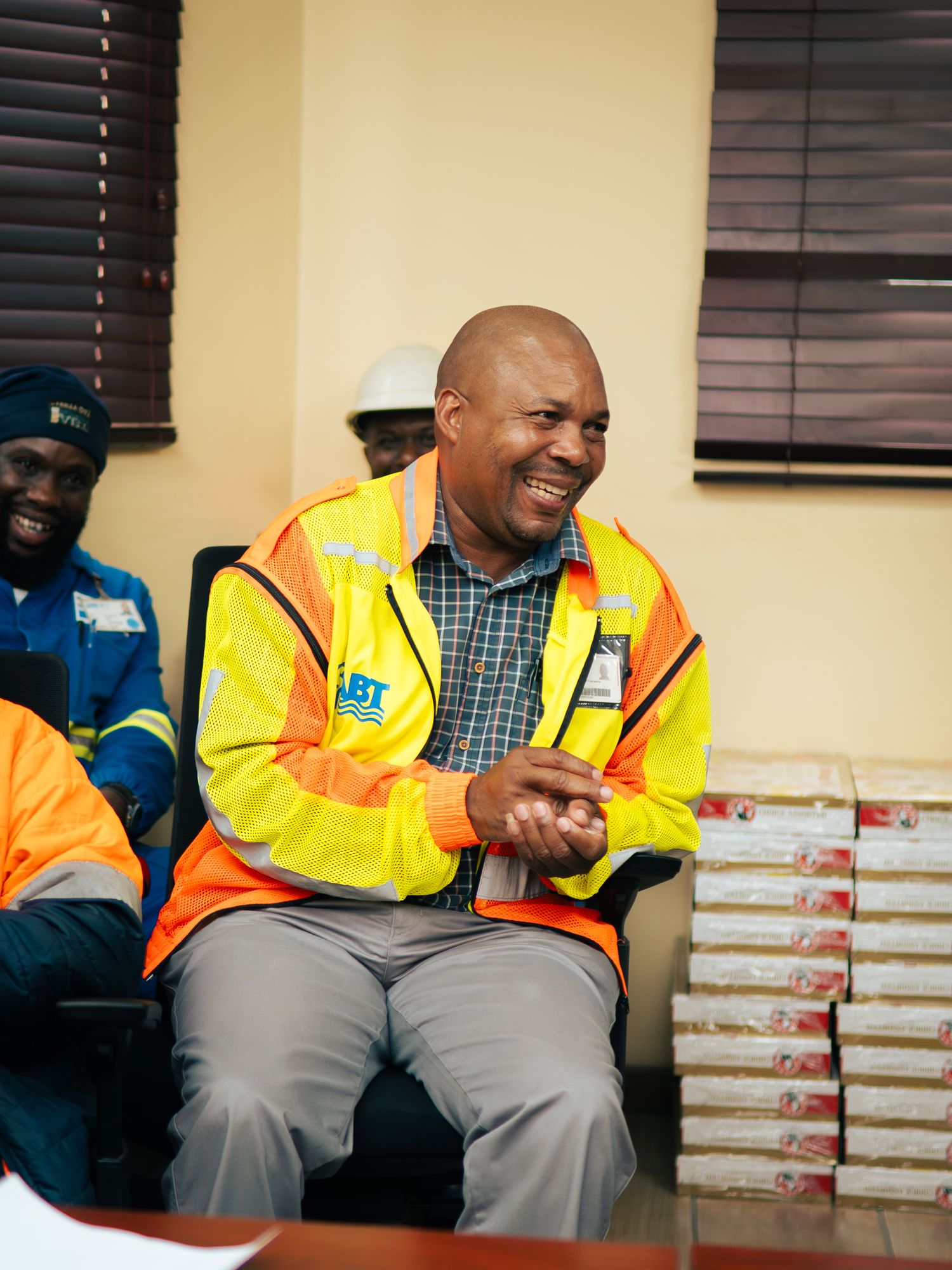 Smiling man in yellow safety vest and hard hat sits in office chair among colleagues at a workplace meeting.