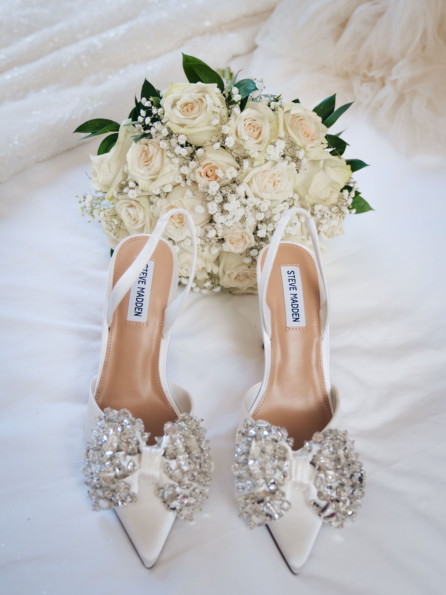 Steve Madden white satin bridal heels with crystal embellishments beside a white rose and baby's breath wedding bouquet.