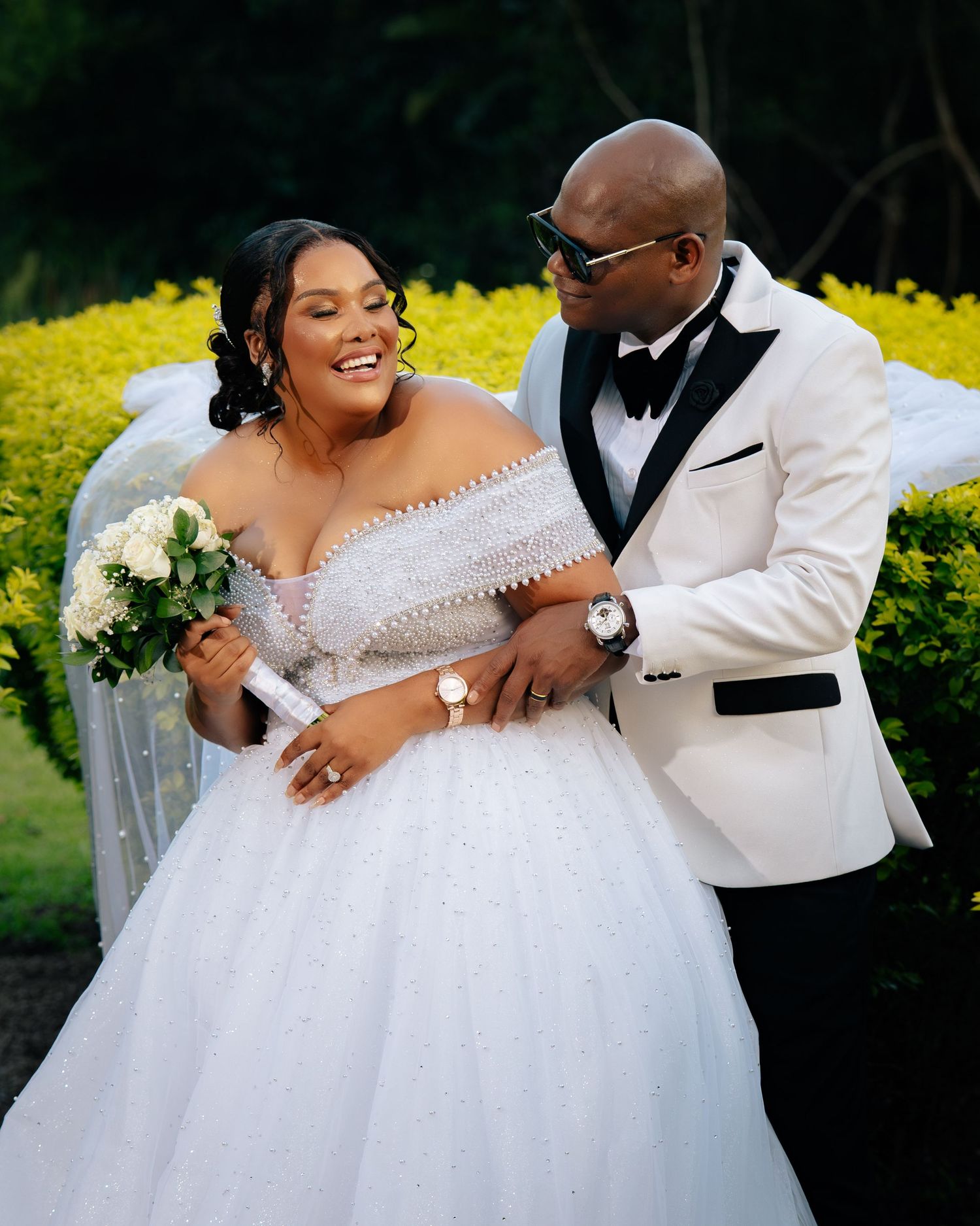 A joyful couple poses outdoors on their wedding day, bride in a sparkling off-shoulder ballgown holding white roses.