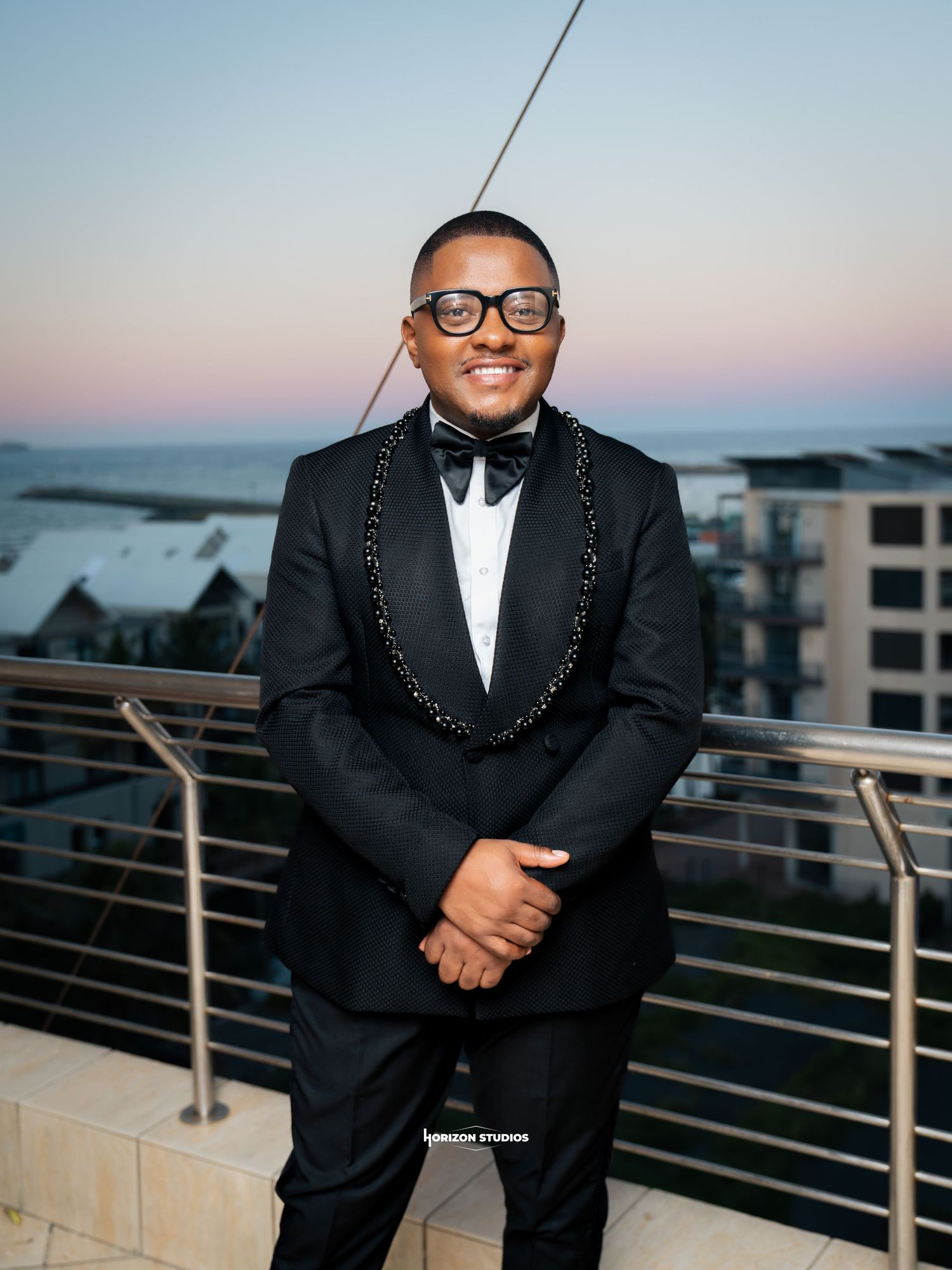 Man in black tuxedo with bow tie smiling on rooftop terrace at sunset with ocean view backdrop.