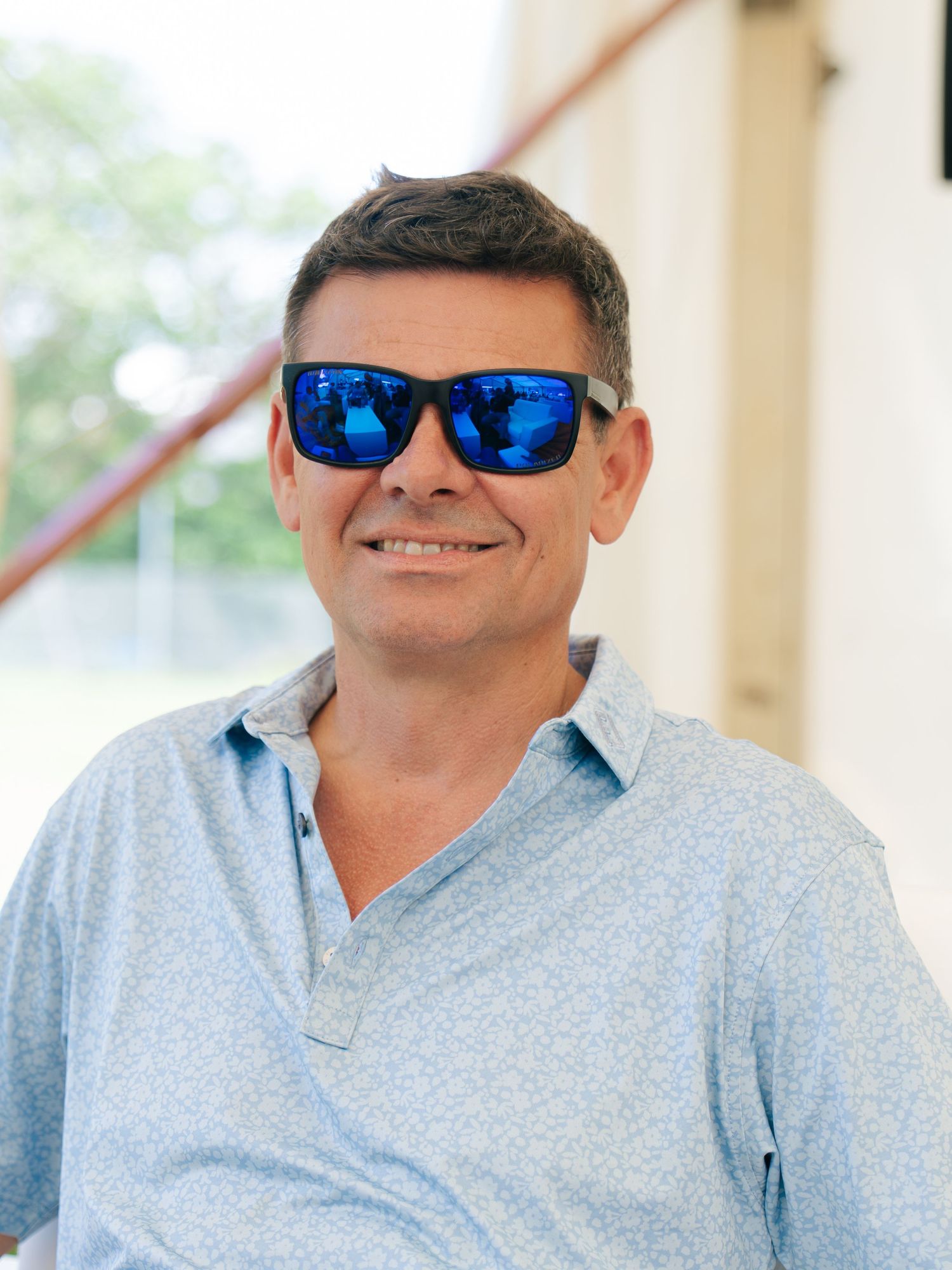 Smiling middle-aged man wearing blue mirrored sunglasses and a light blue polo shirt outdoors.