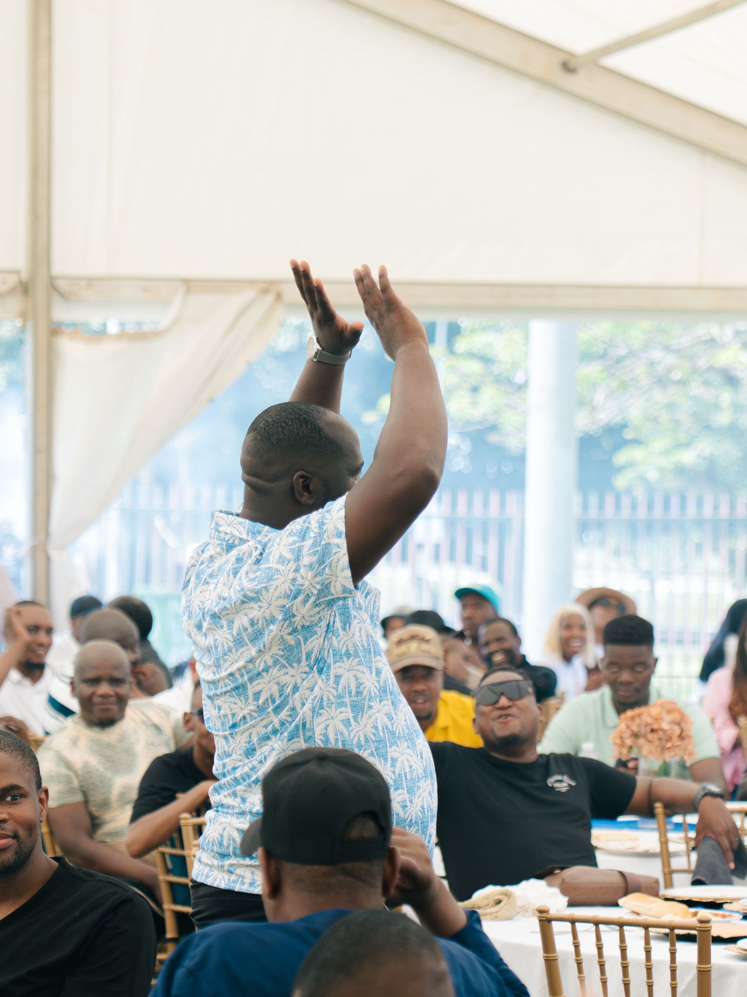 Man raises hands at crowded outdoor event under white tent with guests seated at tables.