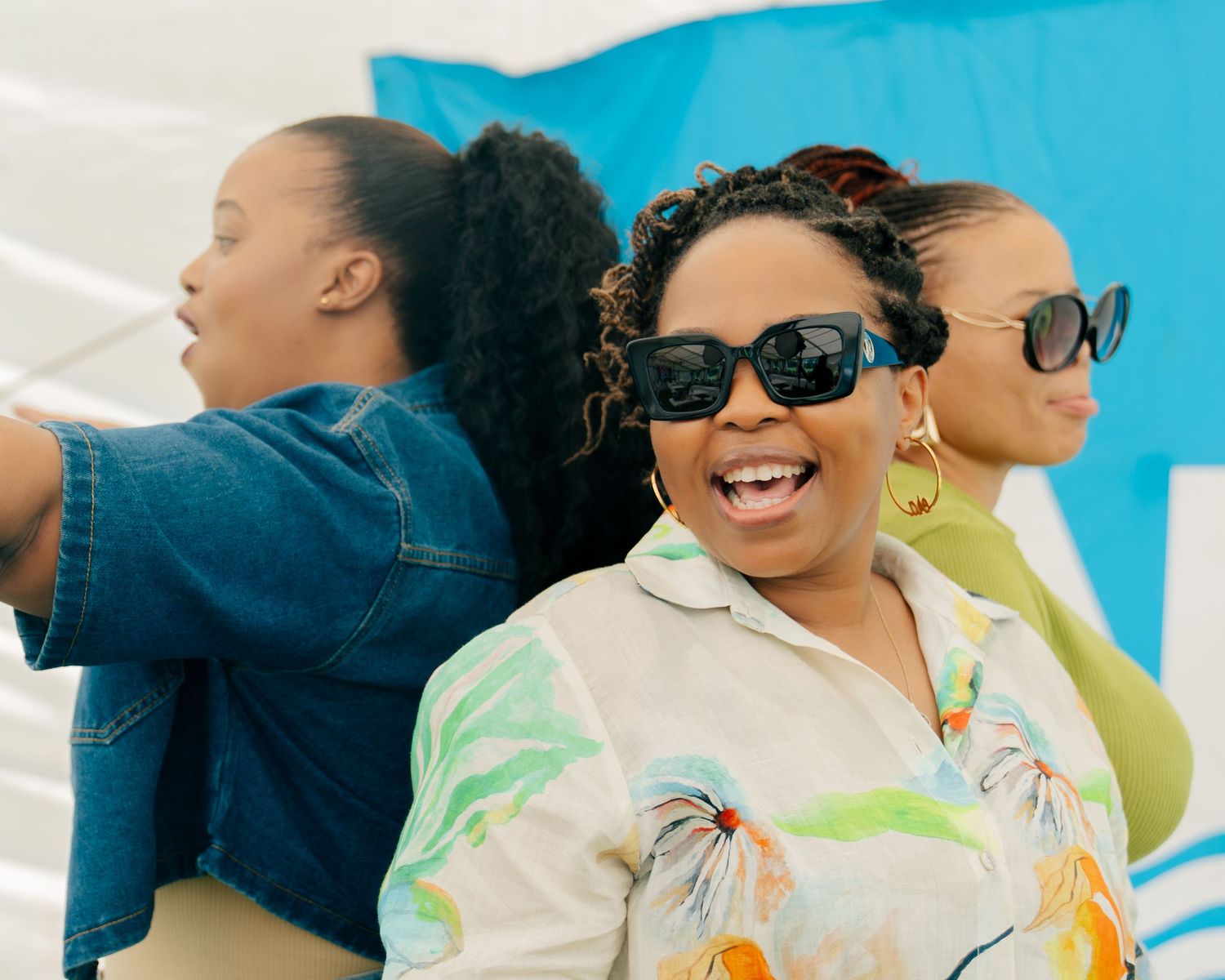 Three women posing outdoors, smiling and wearing sunglasses, dressed in colorful and casual summer outfits.