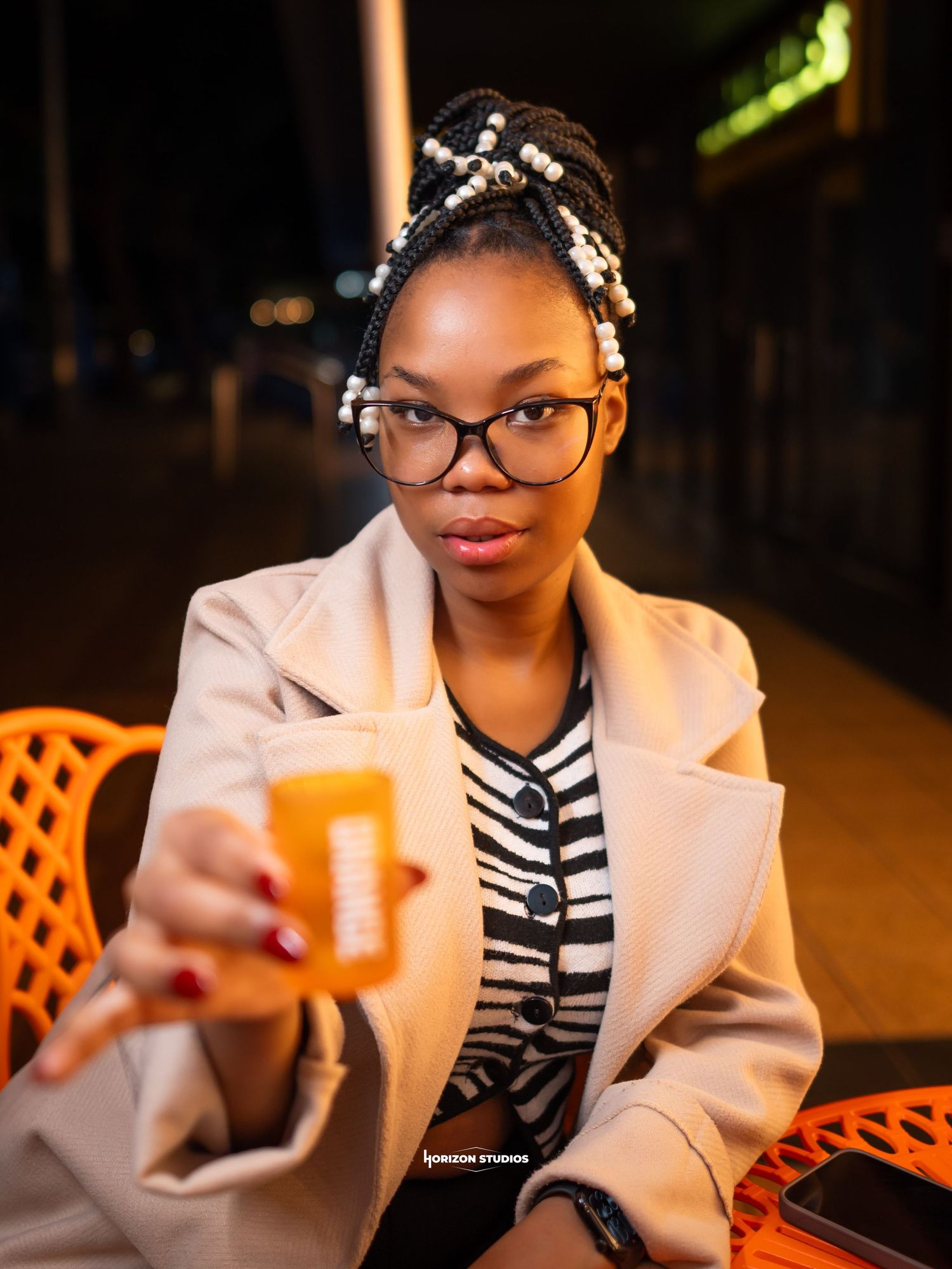 Young Black woman with braided updo and glasses holds an orange drink at an outdoor cafe table at night.