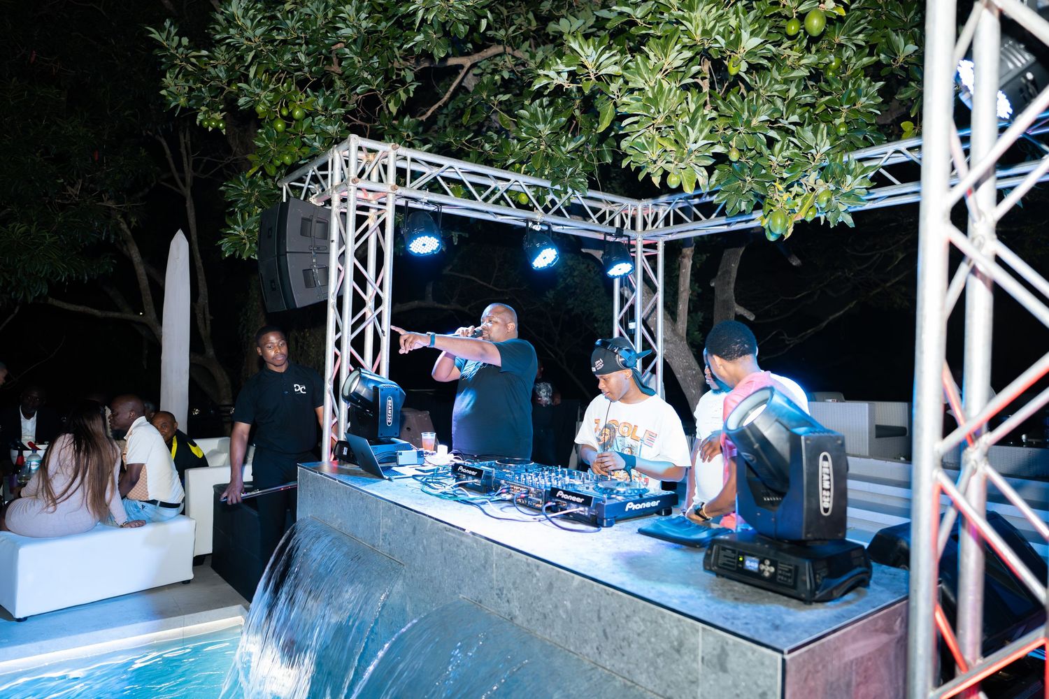 DJs performing at an outdoor nighttime party event with stage lighting and poolside lounge area.