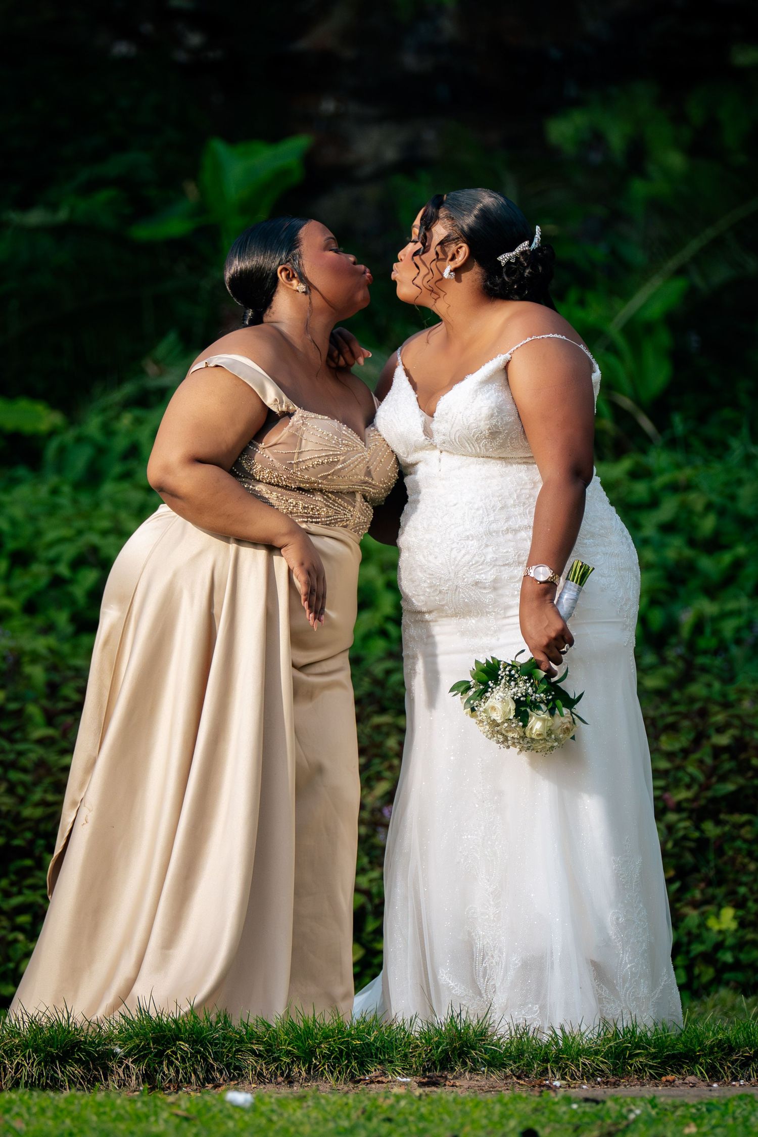 Two women in formal gowns share a tender moment, one in white bridal dress holding bouquet, outdoors.
