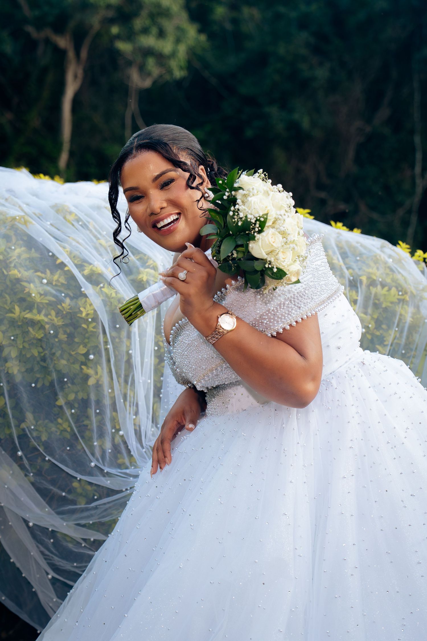Smiling bride in white ballgown holding white flower bouquet with long veil outdoors.