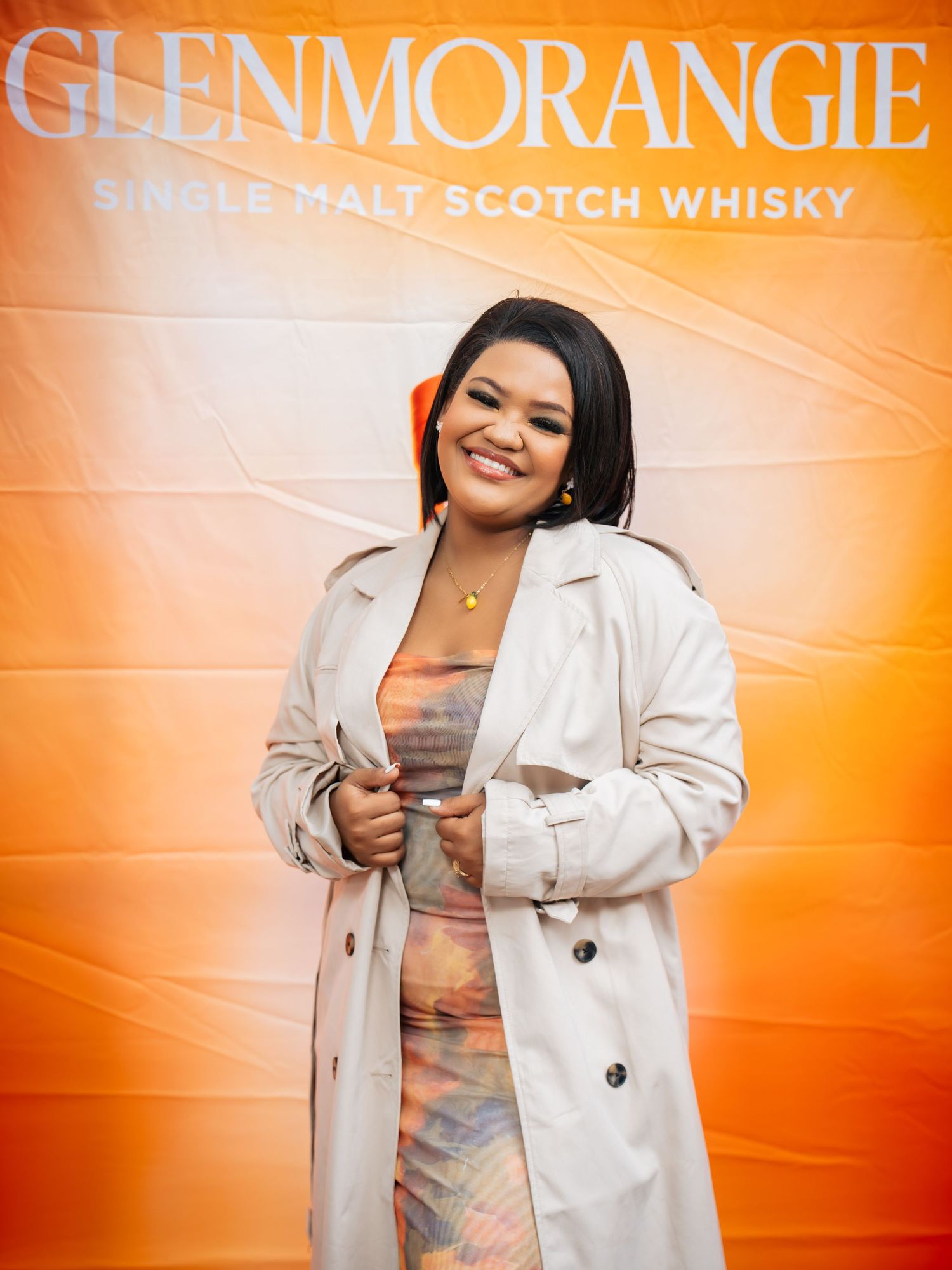 Woman smiling in colorful dress and trench coat at Glenmorangie Single Malt Scotch Whisky event.