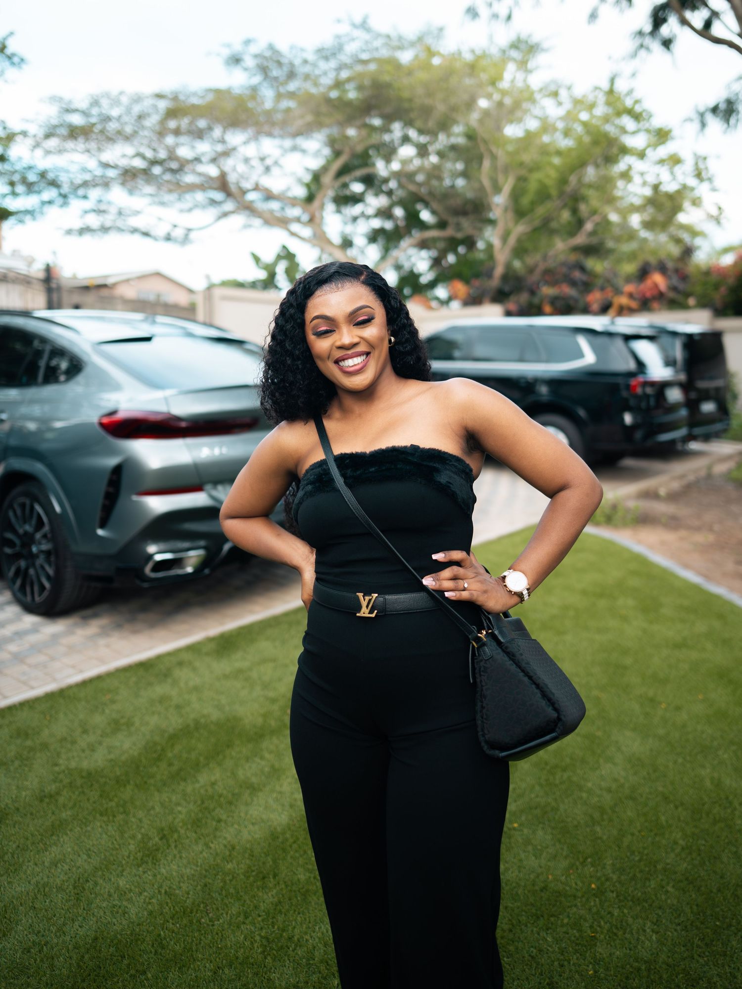 Woman in black strapless jumpsuit with Louis Vuitton belt bag smiling outdoors near luxury cars.