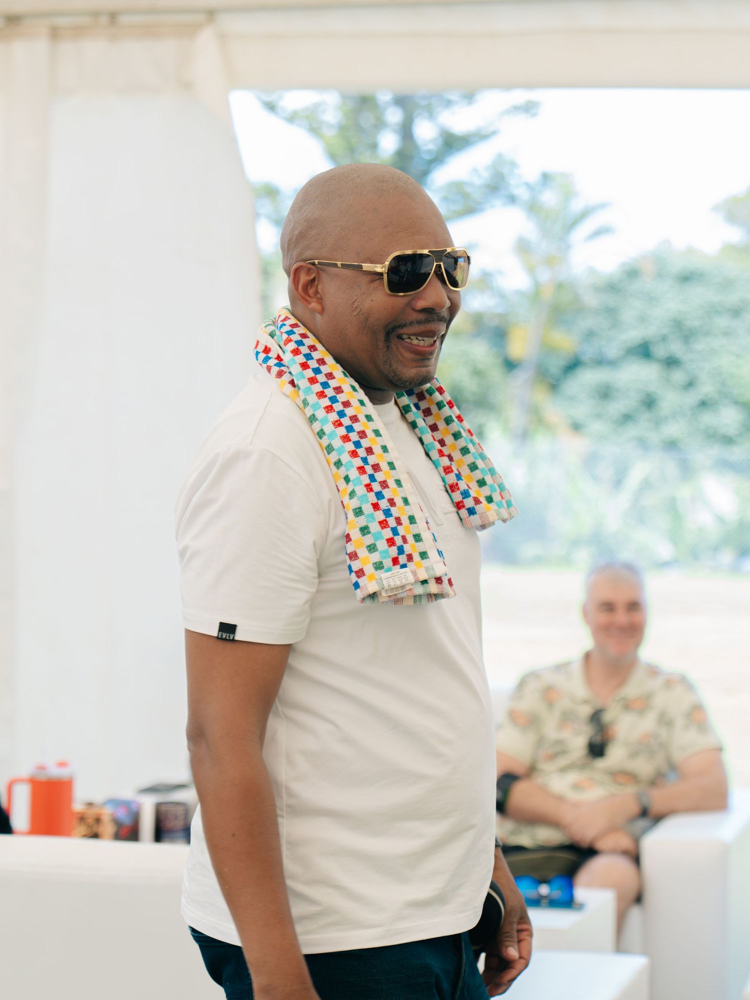 Bald man wearing sunglasses and colorful scarf smiles in white polo shirt at outdoor event with palm trees.