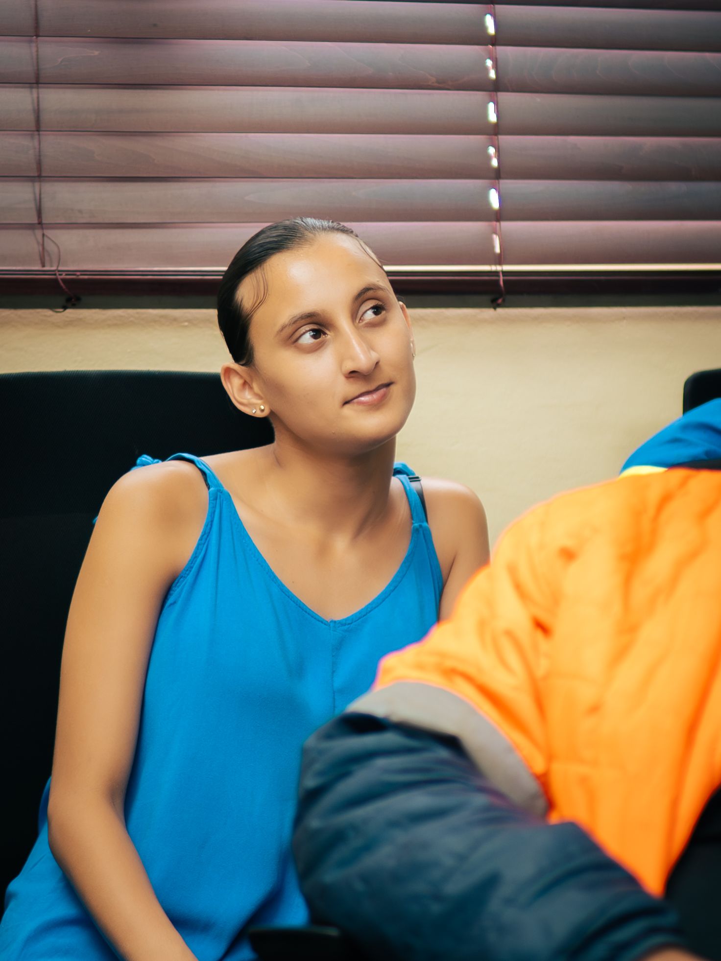 Young woman in blue tank top sitting in chair, looking thoughtful, with blinds in background.