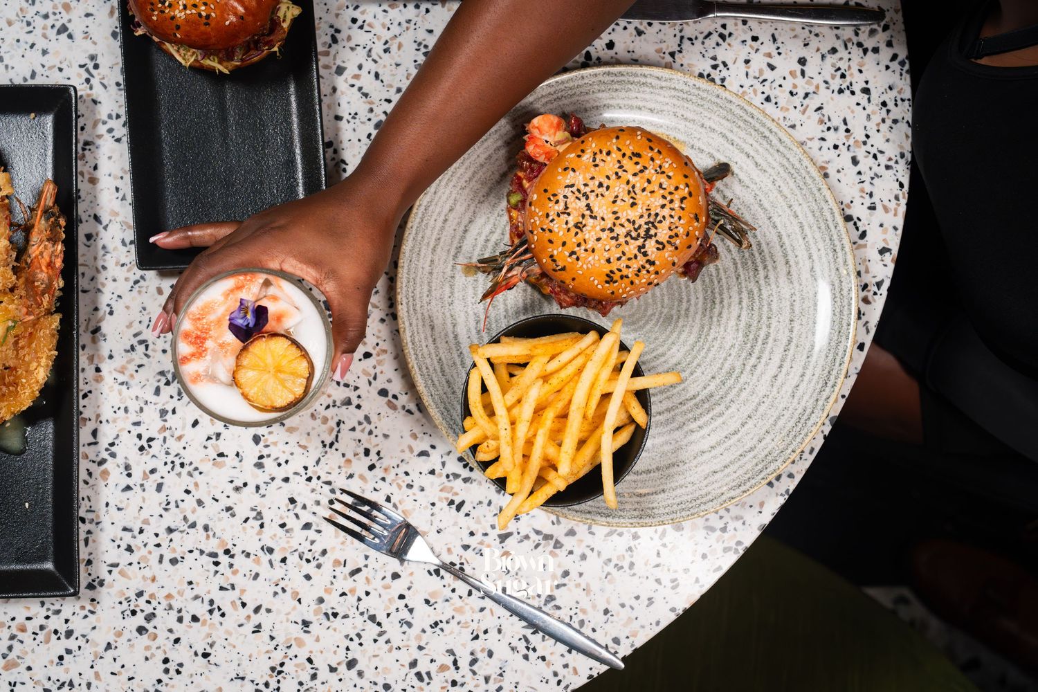 Overhead view of a gourmet burger with fries on a plate, as a hand holds a cocktail on a terrazzo table.