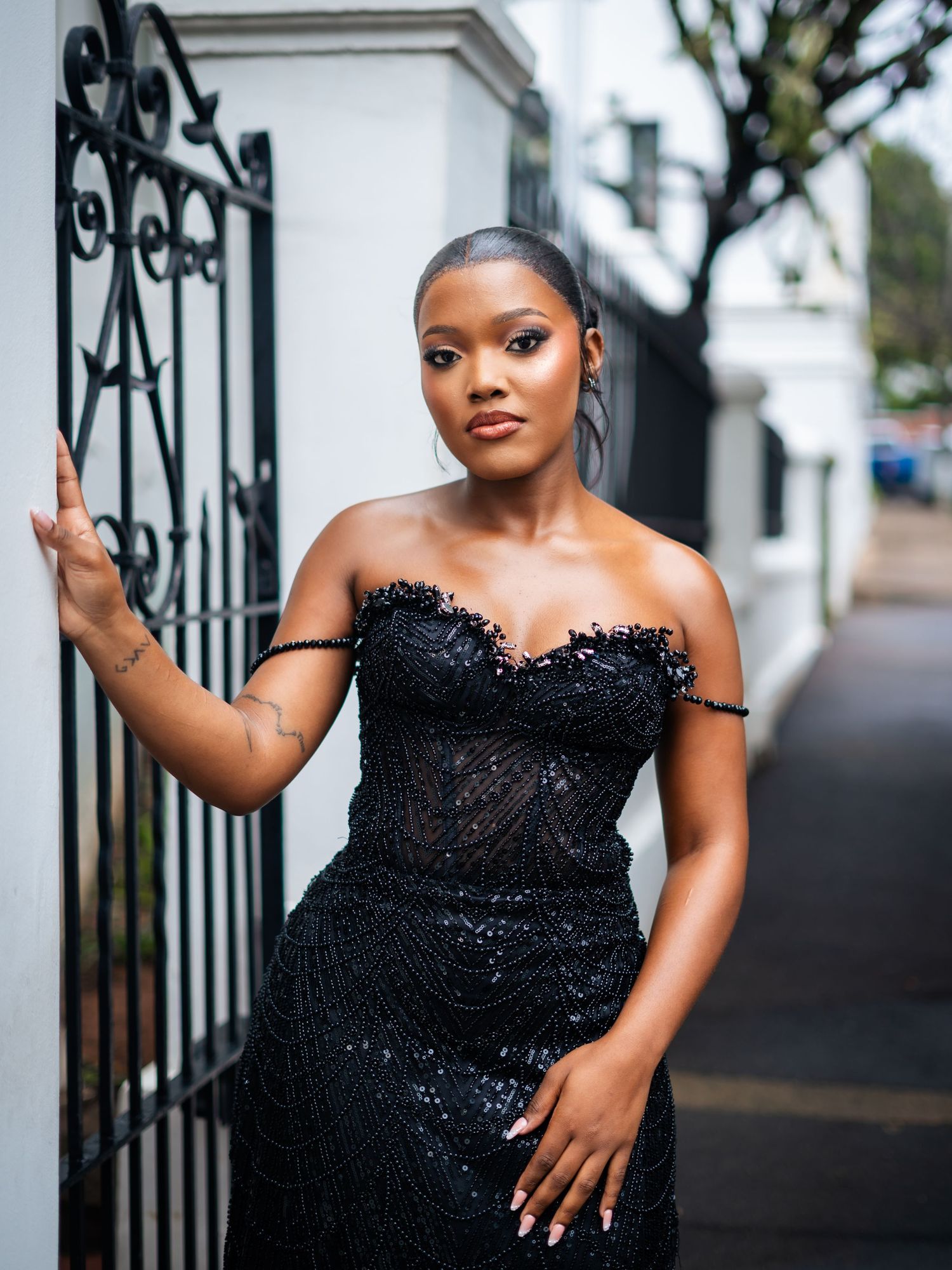 Woman in elegant black sequin off-shoulder gown posing gracefully by ornate iron gate on a stylish street.