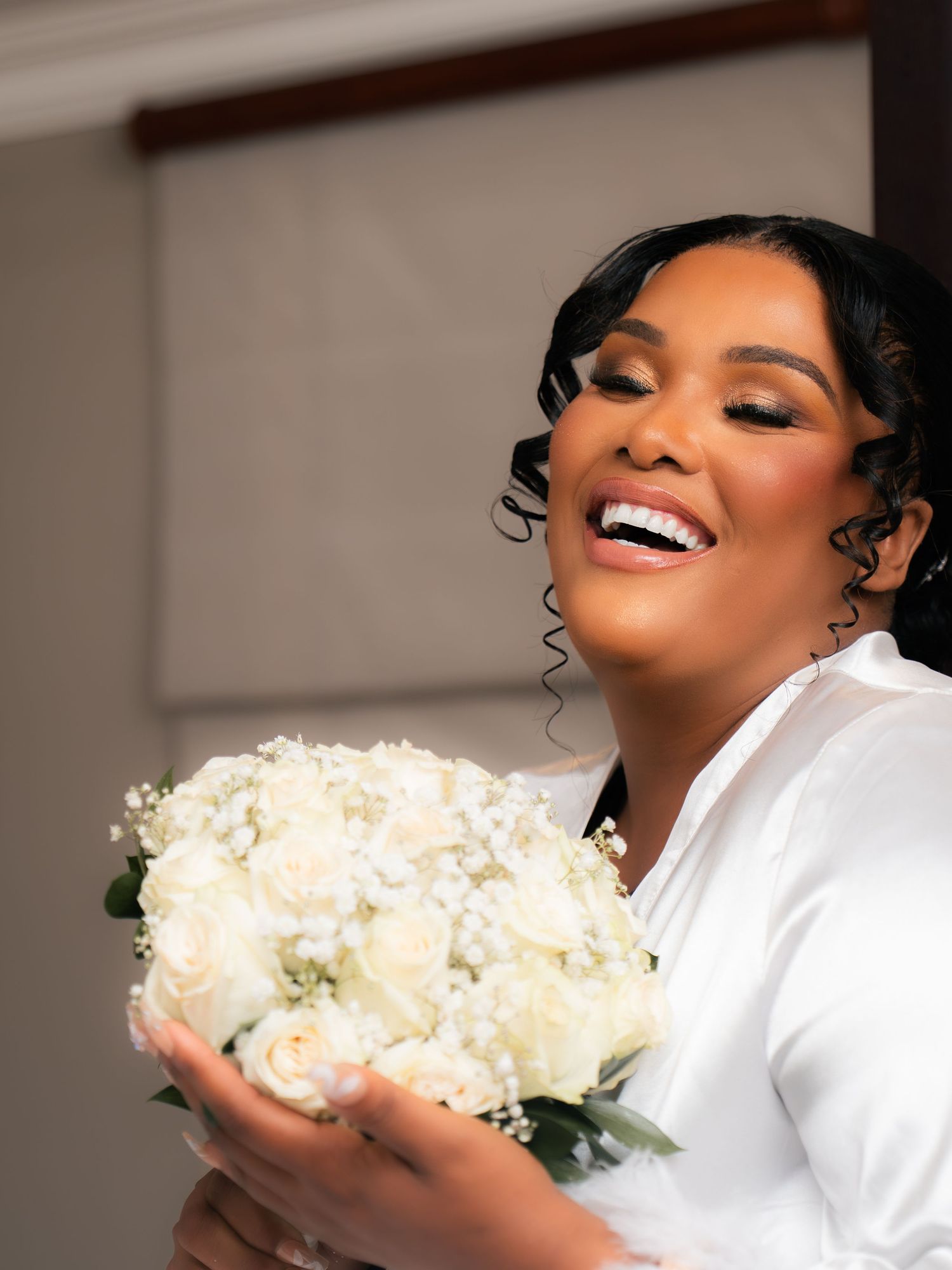 A radiant bride in a white robe smiles joyfully while holding a bouquet of white roses and baby's breath.
