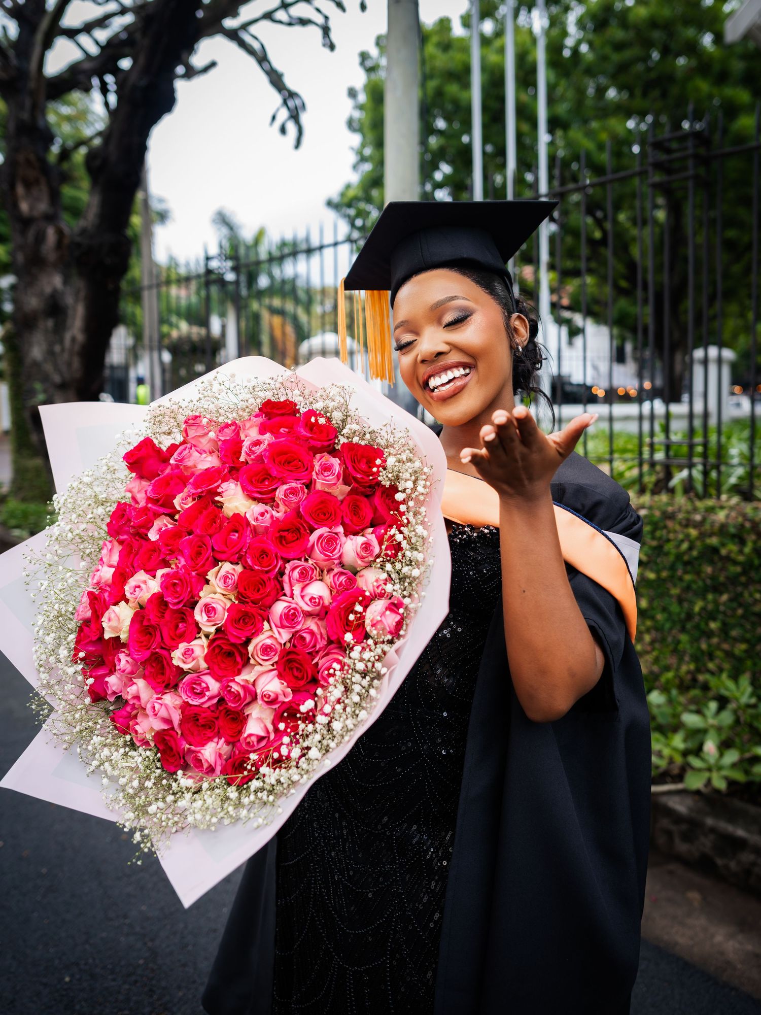 Smiling graduate in cap and gown holding large bouquet of pink and red roses with baby's breath outdoors.
