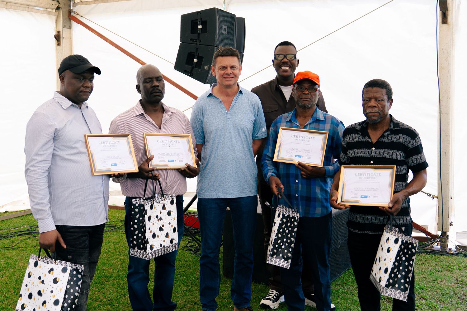 Group of six people posing with certificates and gift bags at an outdoor awards ceremony under a white tent.